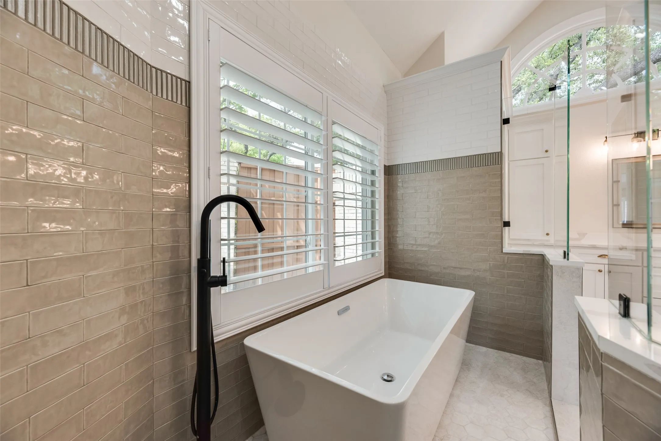 Freestanding soaking tub with a floor-mounted matte black faucet, plantation shutters, and a frameless glass shower enclosure