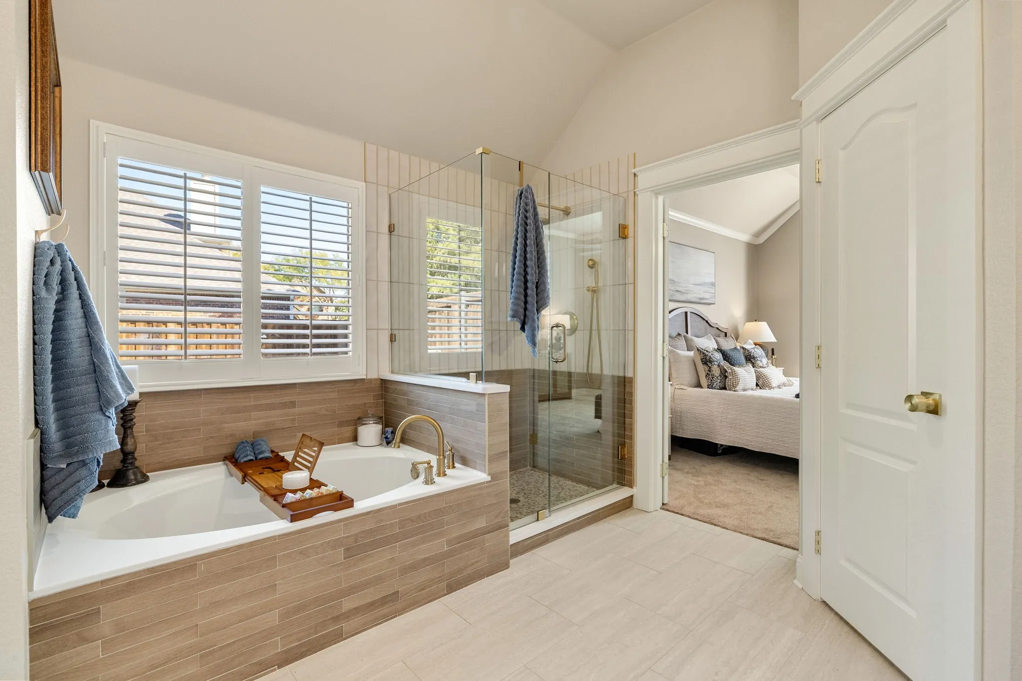 Soaking tub with brass-tone fixtures, wood-finish tile surround, and plantation shutters