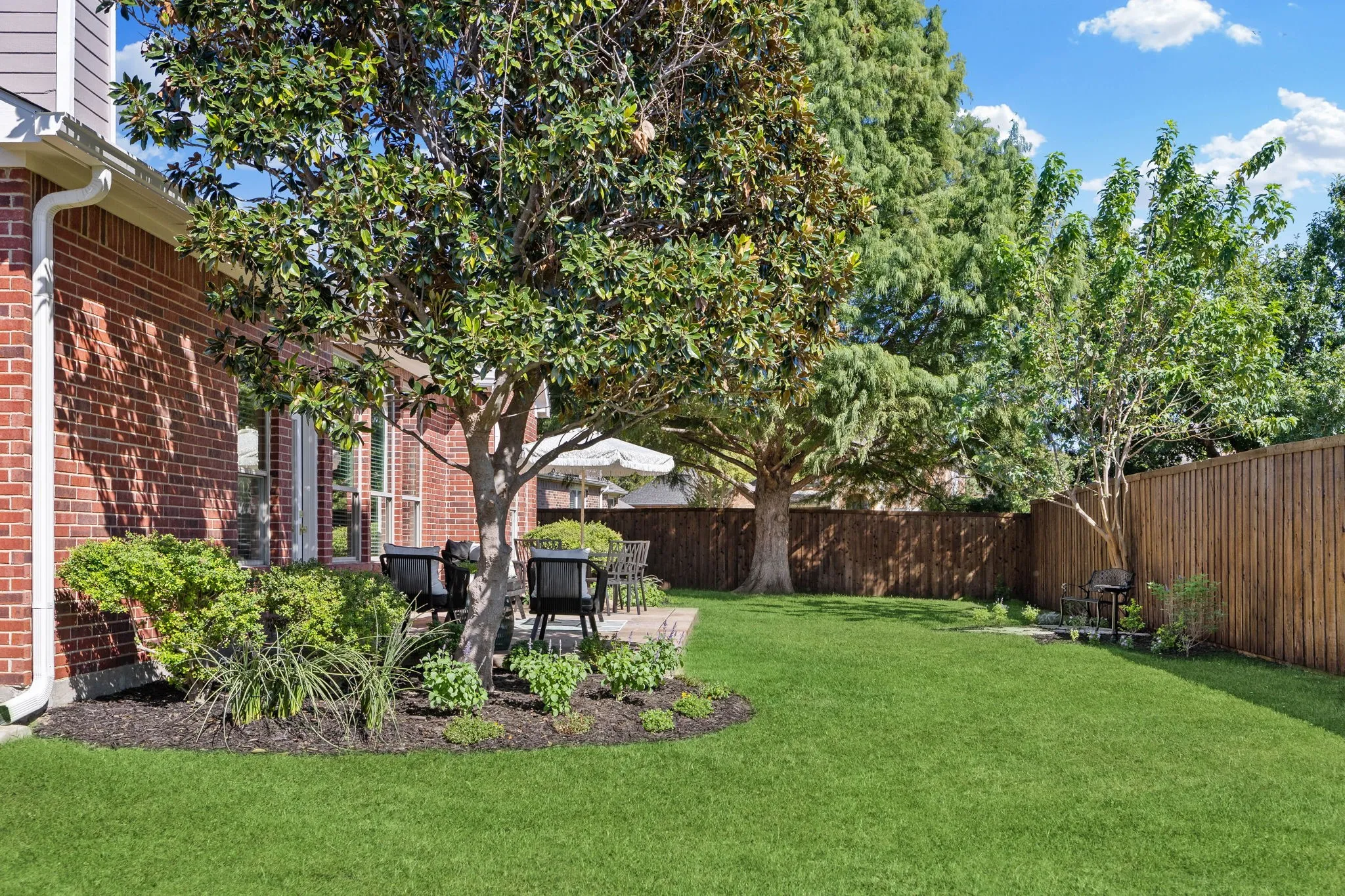 Brick exterior with large windows, mature trees, wood fence, and a patio area