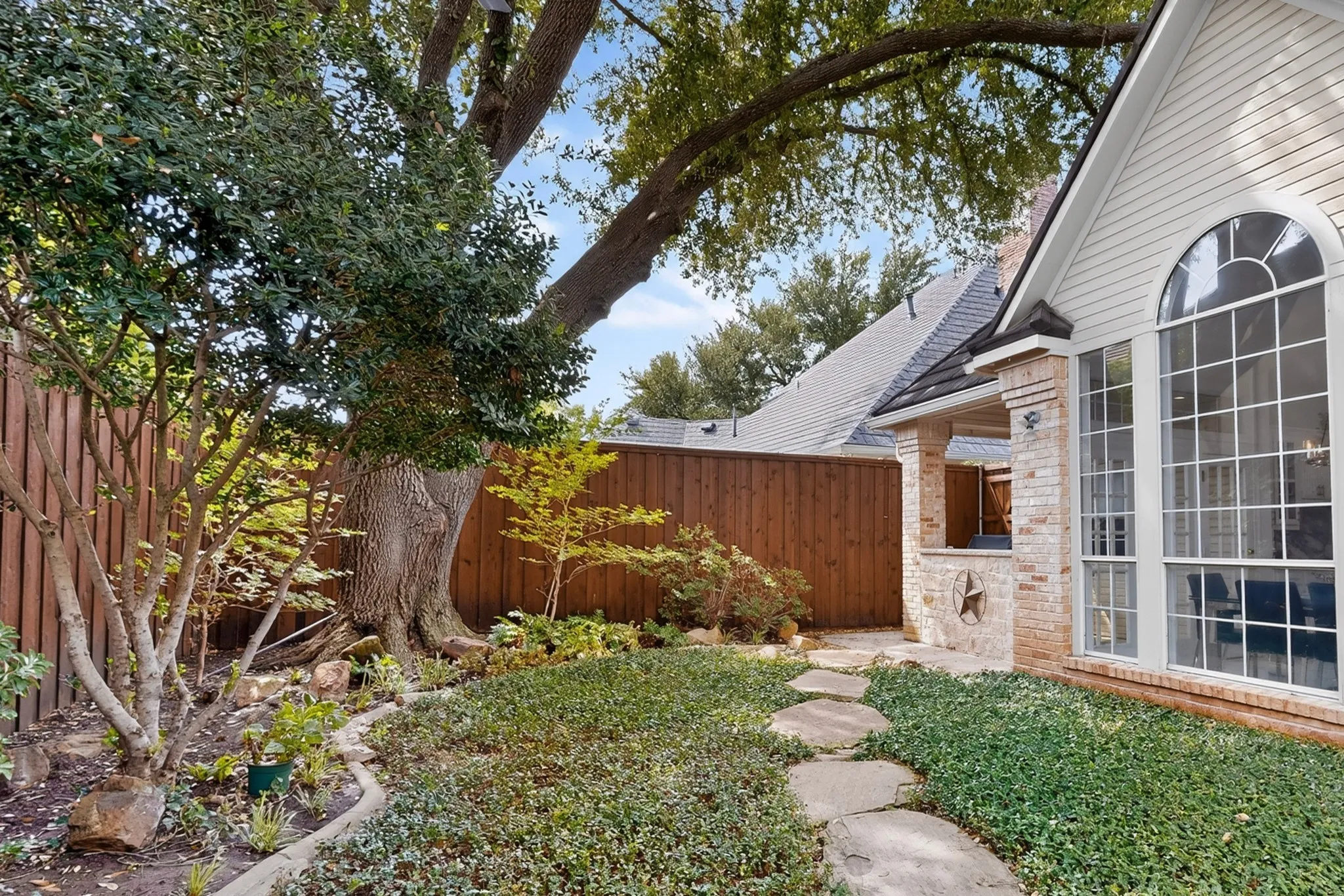 Manicured landscaping with mature trees, stone pathway, brick exterior, arched window, and covered patio