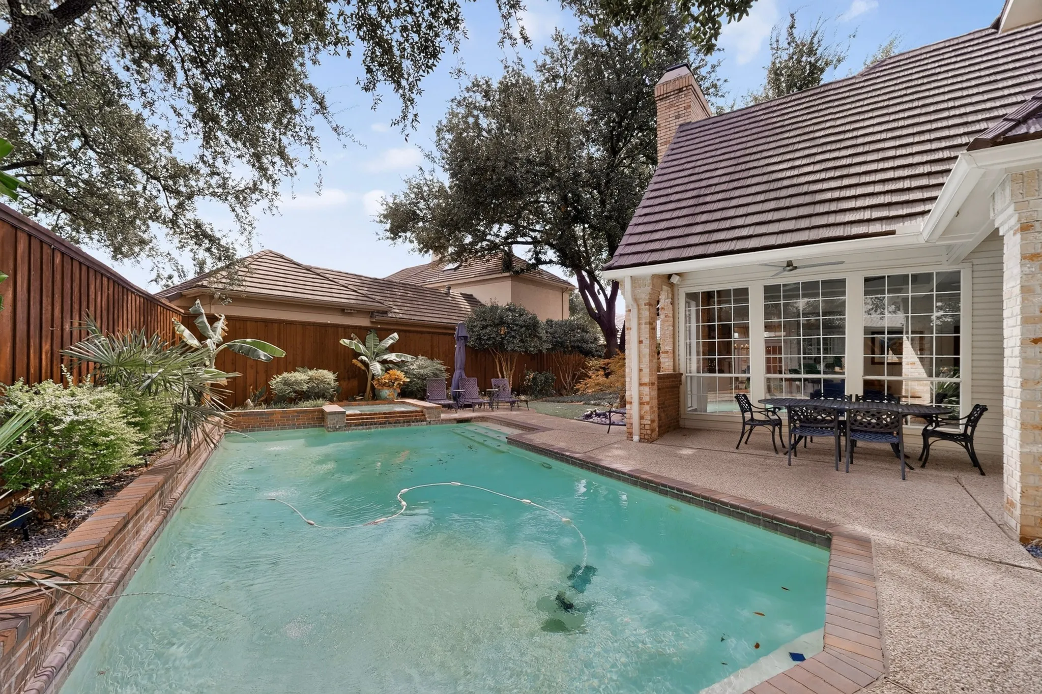 Resort-style backyard oasis featuring a sparkling turquoise swimming pool with surrounding brick decking, mature shade trees, a covered patio with French doors, and a brick chimney detail on the main residence
