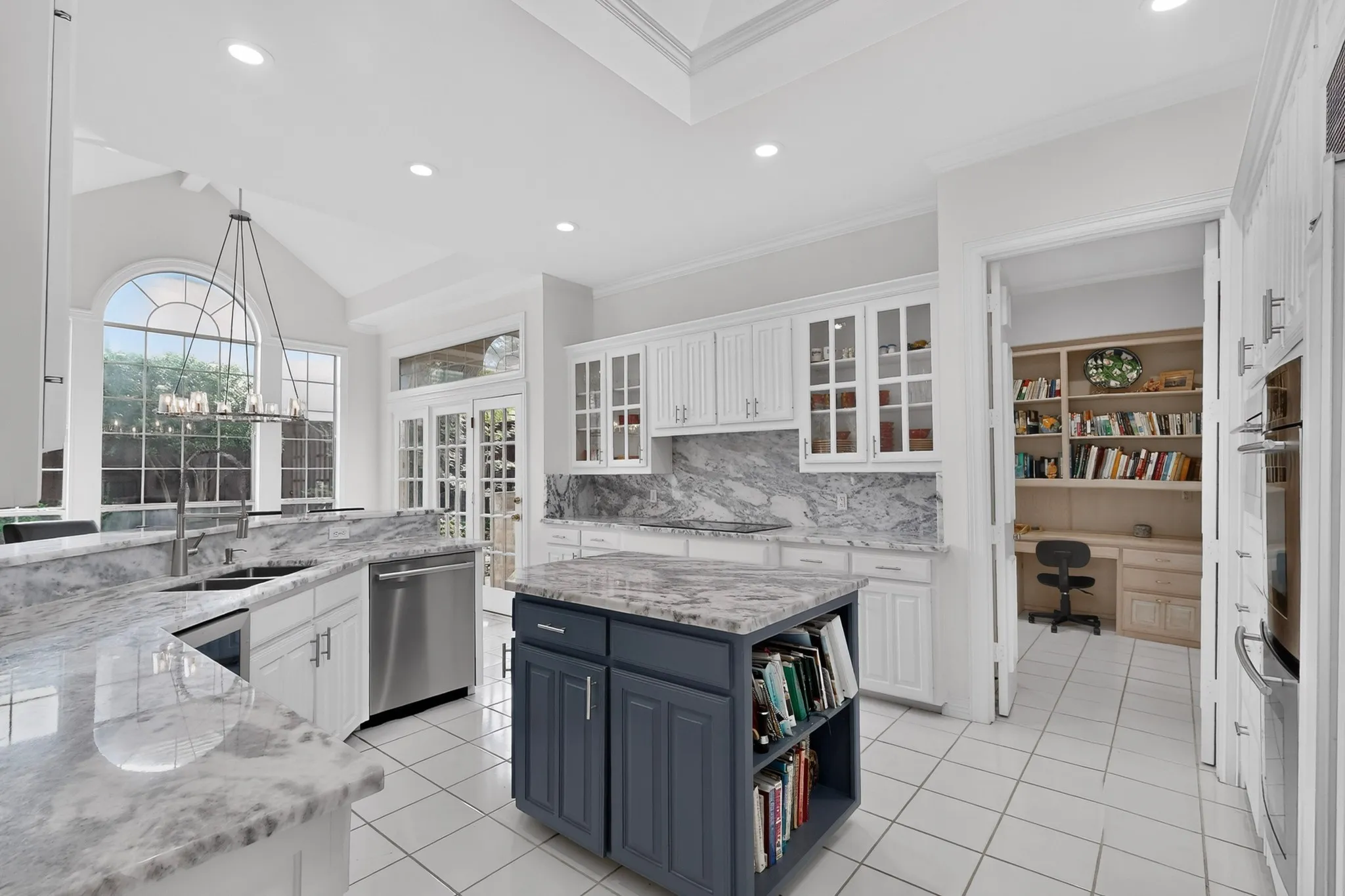 Gourmet kitchen featuring white cabinetry, stone countertops, a central island with built-in shelving, stainless steel appliances, and a vaulted ceiling with an arched window