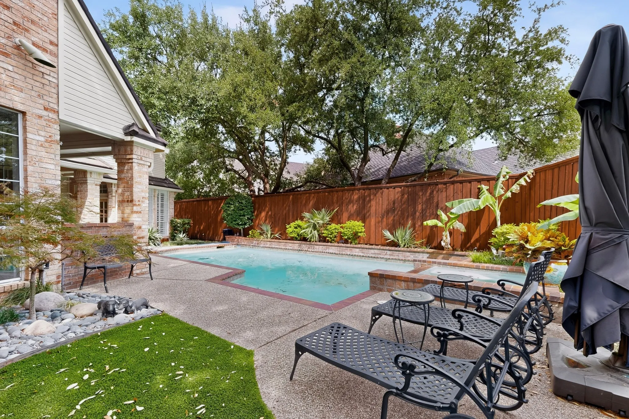 Expansive backyard oasis featuring a resort-style swimming pool with sun lounging area, mature shade trees, manicured lawn, wood privacy fencing, and brick exterior home with covered patio access