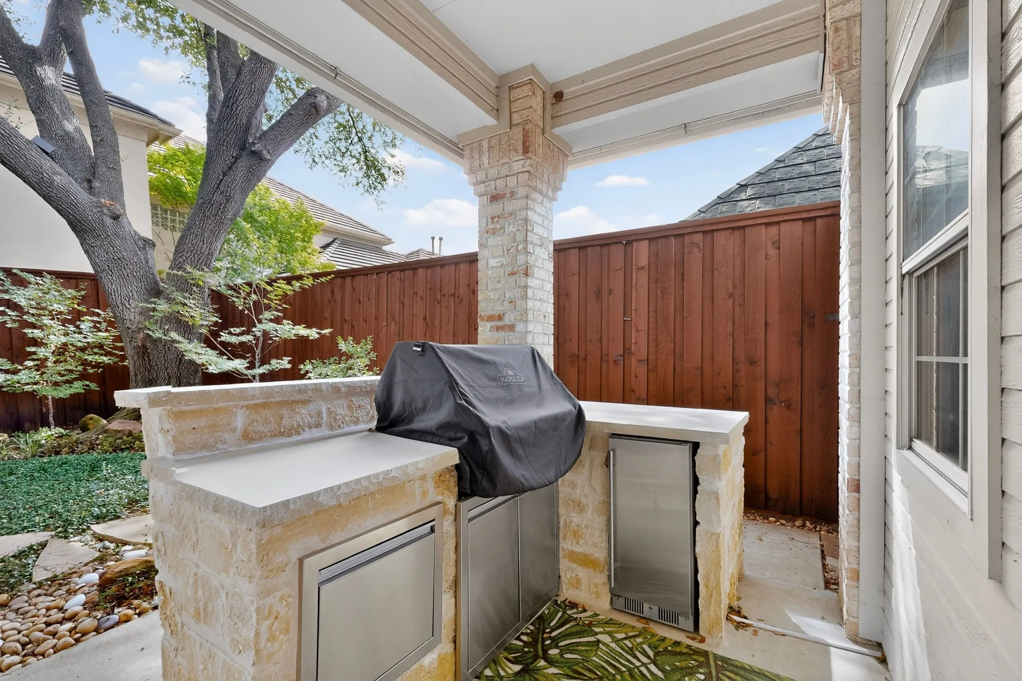 Covered patio featuring a built-in stone grill with stainless steel appliances and concrete countertop, framed by a mature shade tree and ornamental fencing