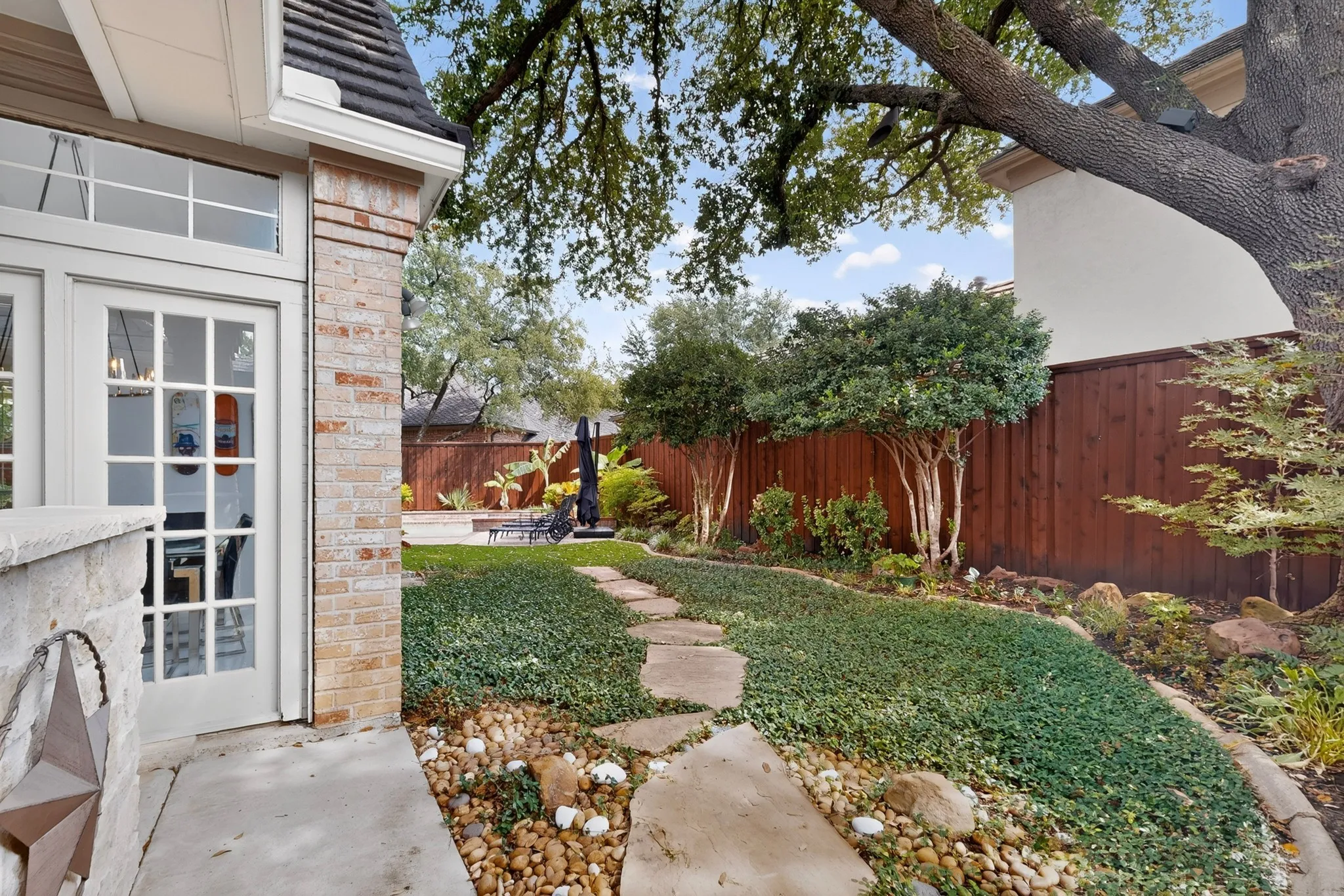 Brick exterior with stone accents, French doors, mature trees, wood fencing, and a stone-paver pathway