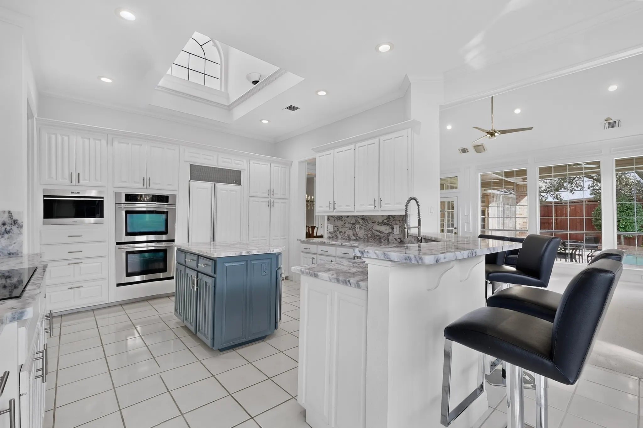 Sophisticated open-concept kitchen and dining area featuring white cabinetry with stainless steel appliances, a dual-island layout with waterfall-edge countertops, tile flooring, recessed and pendant lighting, and direct sightlines to the adjoining living space with large windows providing natural light and outdoor access