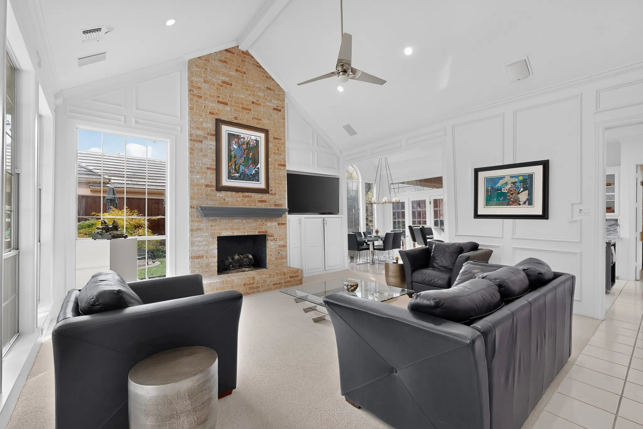Vaulted ceiling with a contemporary ceiling fan, brick fireplace with mantel, built-in cabinetry, large windows, and tile flooring
