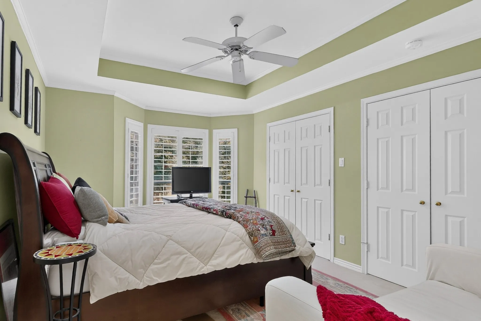 Tray ceiling with a ceiling fan, bay window with plantation shutters, and two sets of double panel doors