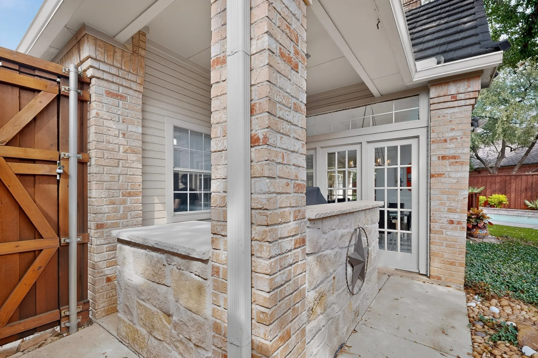 Covered patio with rusticated brick pillars and stone veneer detailing, complemented by a wooden barn-style gate and mature landscaping along the property perimeter