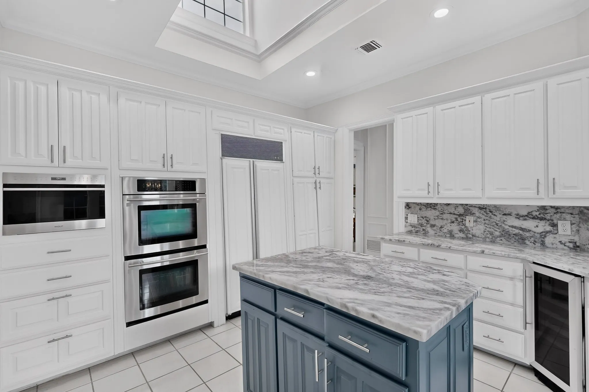 Chef's kitchen featuring white cabinetry with stainless steel hardware, dual wall-mounted ovens, a blue-gray island with marble-finish countertops, stainless steel appliances, tile flooring, and skylight illumination overhead