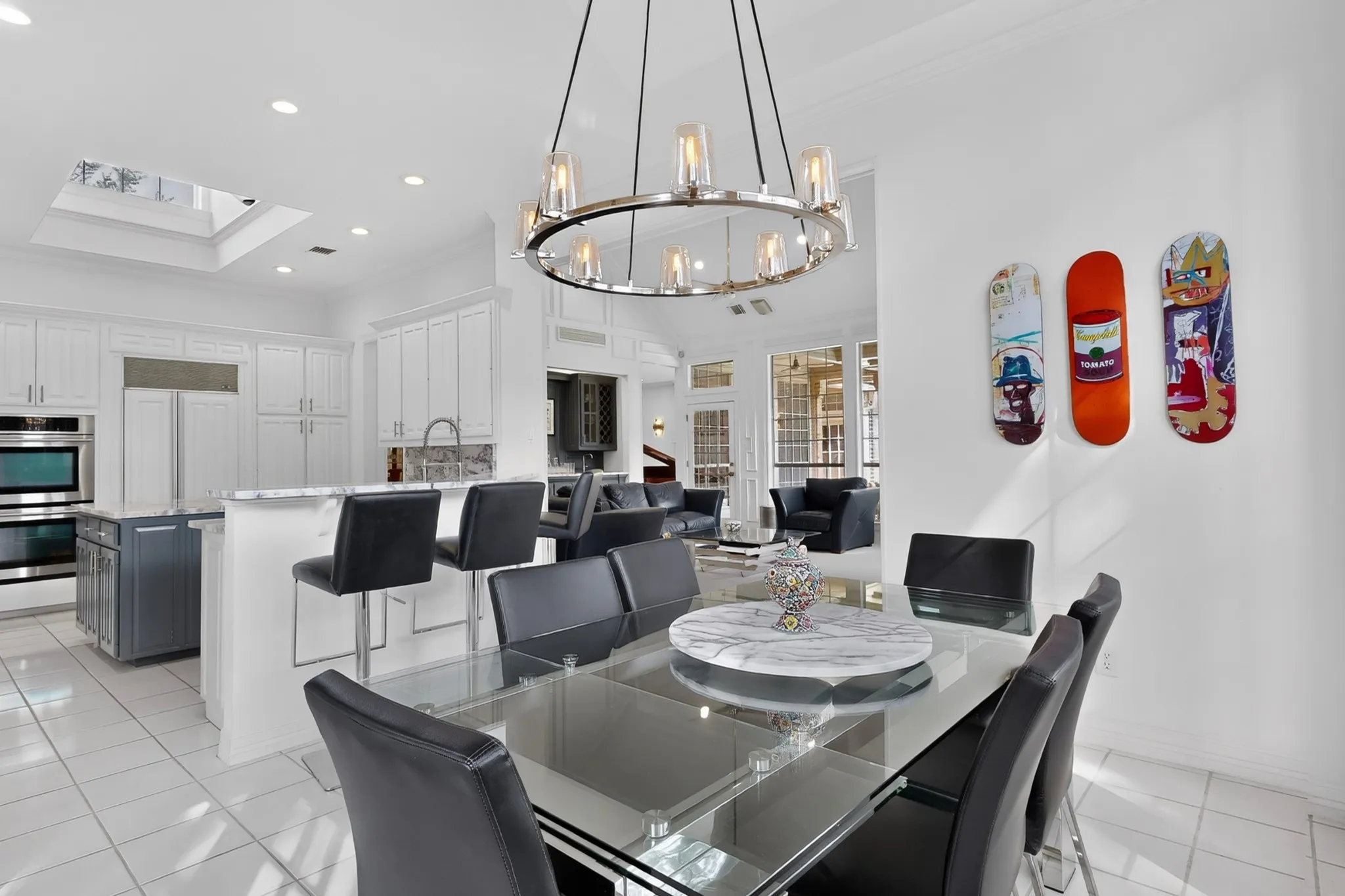 Contemporary dining and kitchen showcase featuring a round glass-top dining table with black upholstered chairs, stainless steel appliances, white cabinetry with integrated refrigeration, light-filled layout with recessed ceiling lighting, and a modern pendant fixture with warm-toned accents