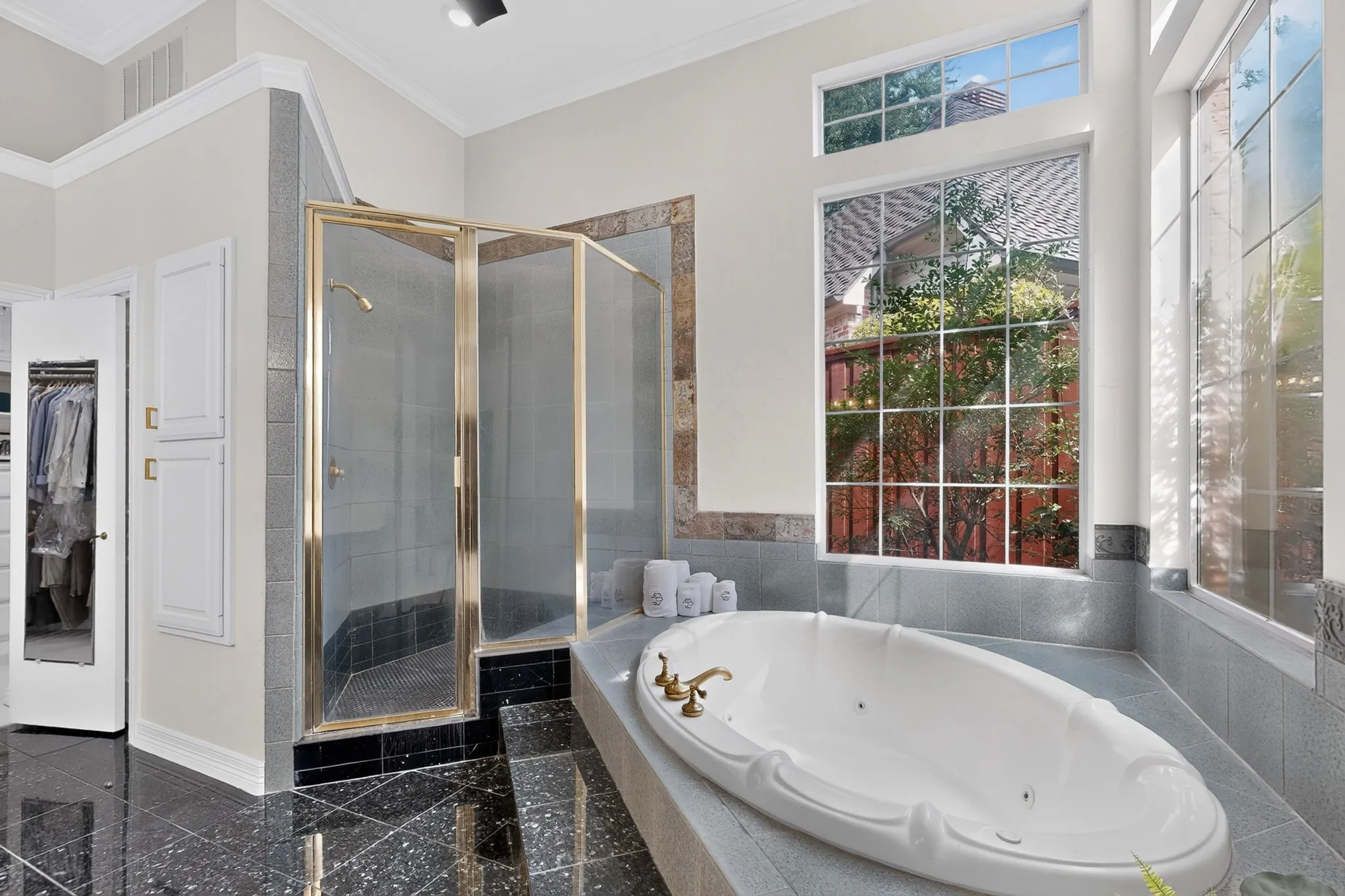 Soaking tub with brass fixtures, glass-enclosed shower, black polished tile flooring, and transom windows