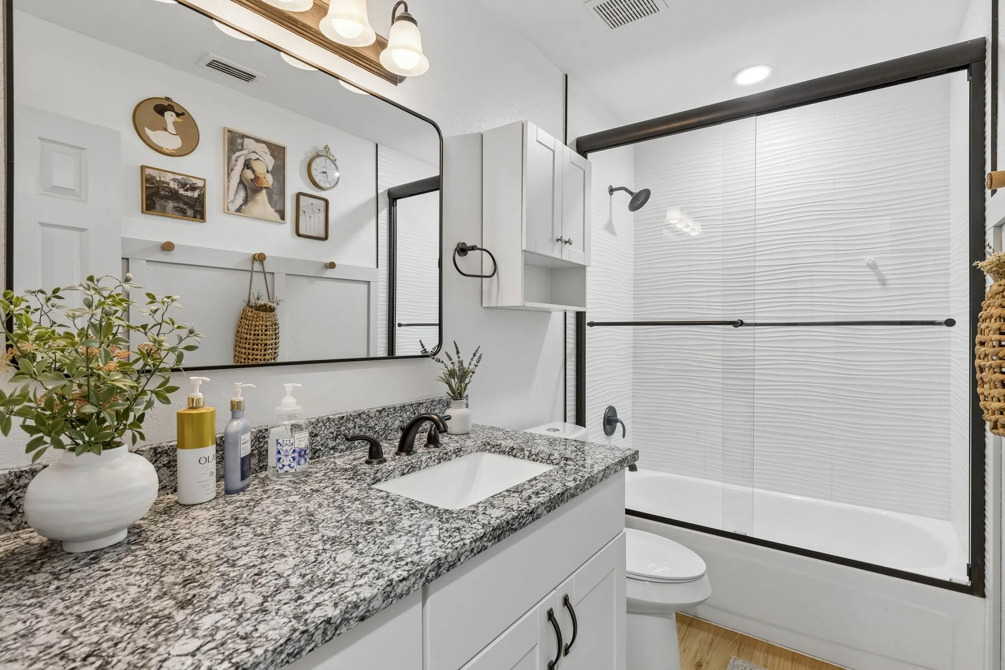 The secondary bathroom displays a large tub in shower with wavy tile, granite counter tops, a framed mirror LVP flooring, recessed lighting, and ample counter and cabinet space.