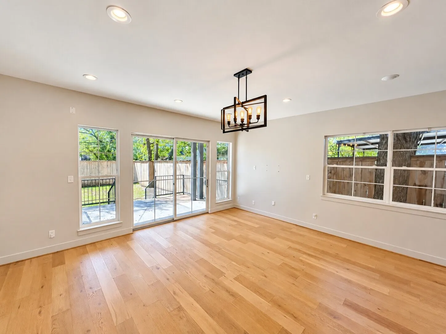 Spacious open room showcasing gleaming wood-finish flooring throughout, abundant natural light from multiple glass doors and windows, recessed ceiling lighting, and a contemporary pendant fixture overhead