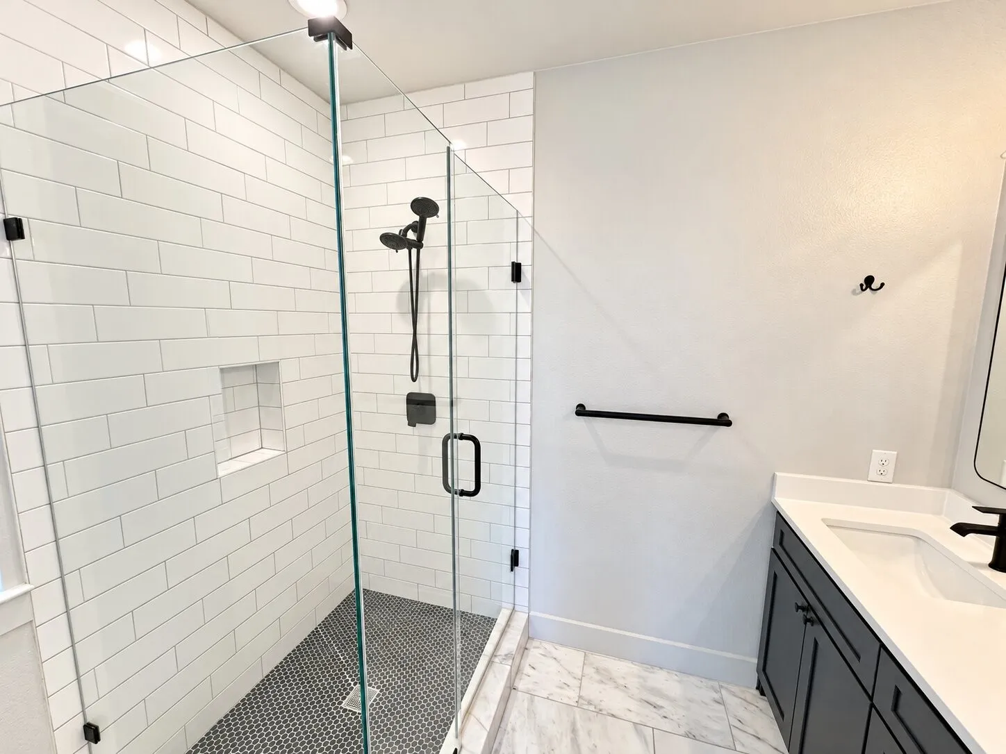 Glass-enclosed shower with white subway tile surround and black penny tile flooring, featuring a built-in shower niche and matte black fixtures