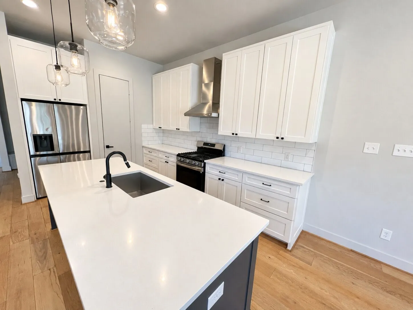 Contemporary kitchen featuring a large center island with an undermount sink, stainless steel appliances, white shaker cabinetry, white subway tile backsplash, and light wood-finish flooring