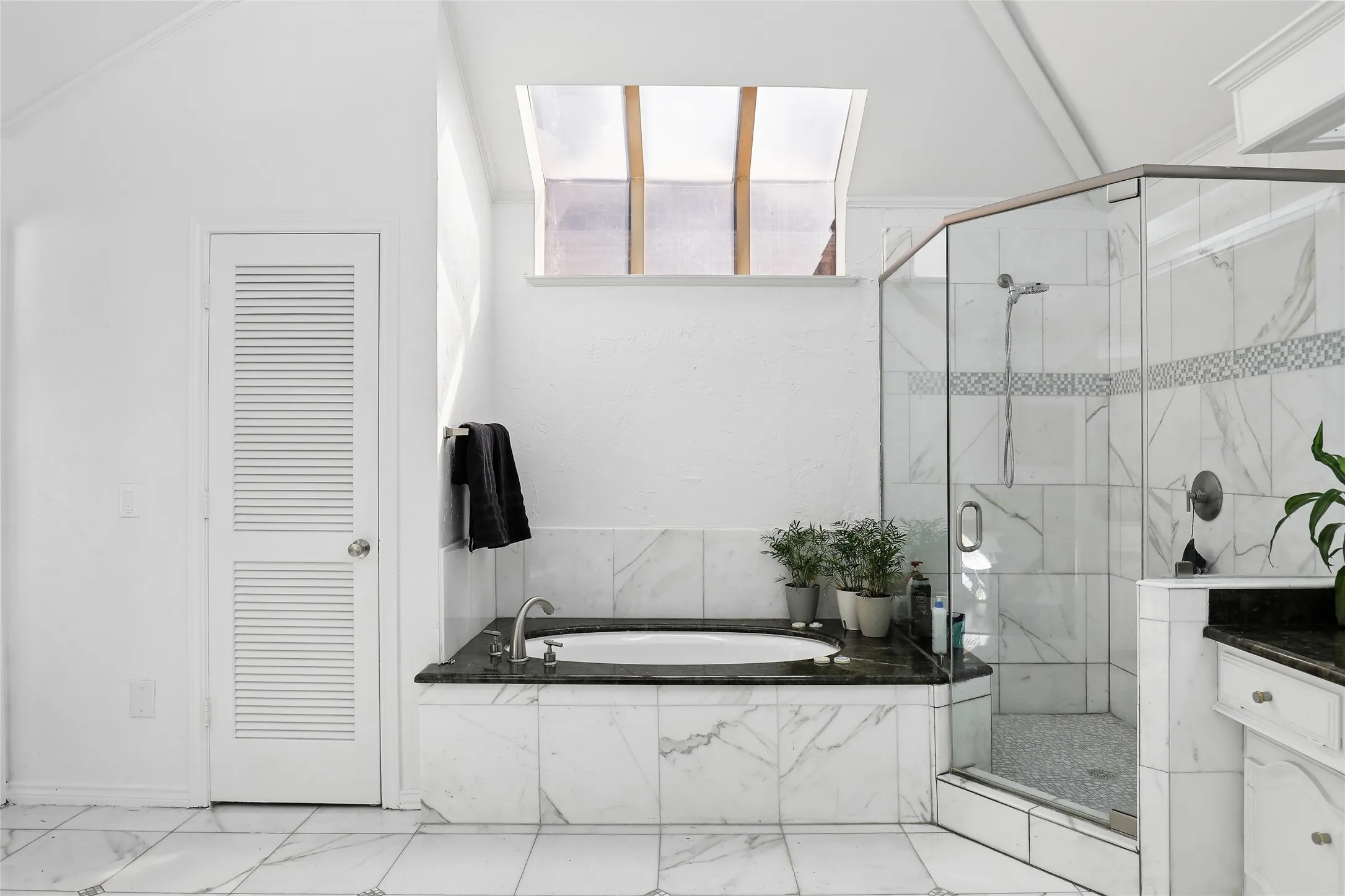 Recessed bathtub with dark stone surround, walk-in shower with clear glass enclosure, white marble-style wall and floor tiling, and a ceiling skylight