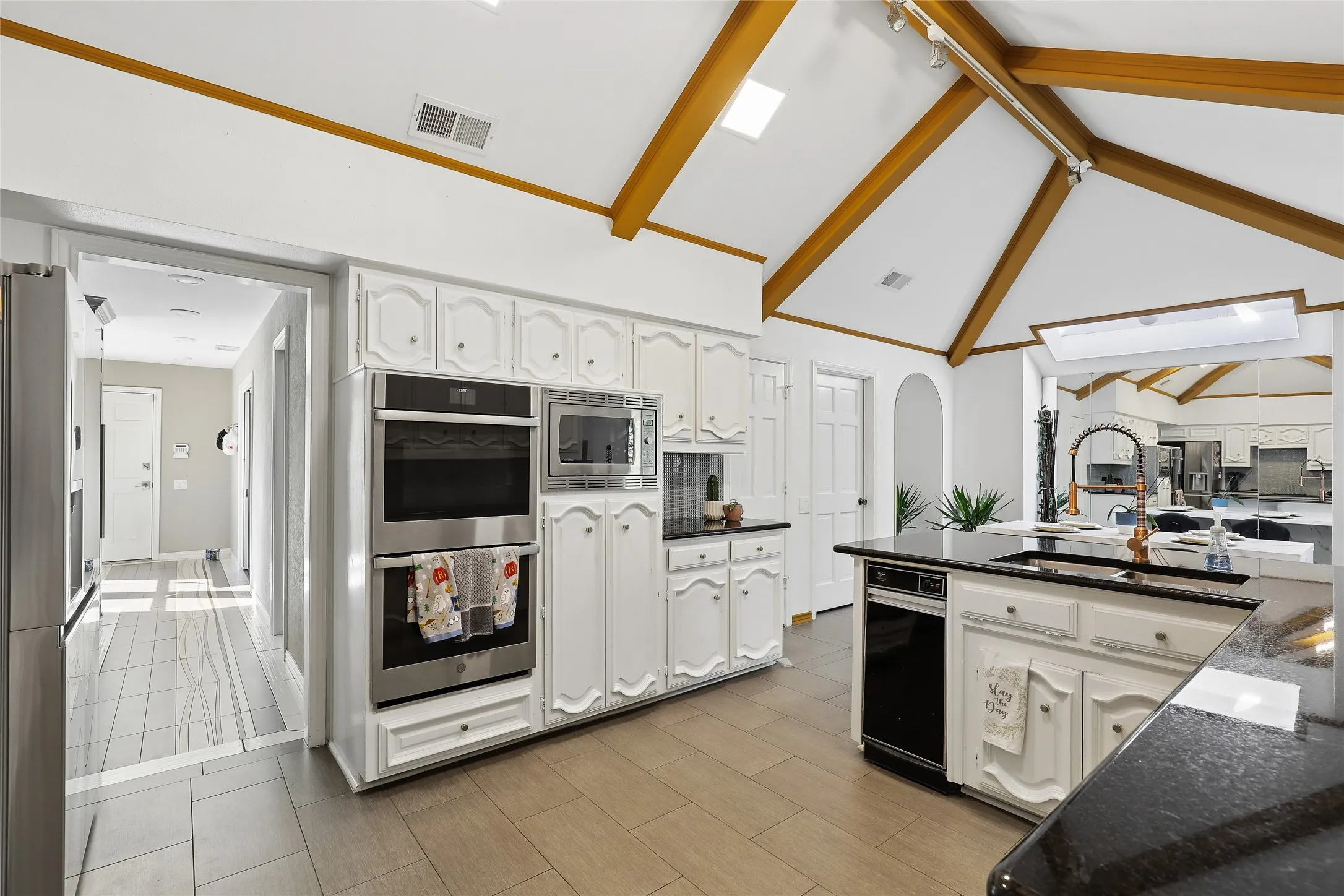 Vaulted ceiling with exposed wood beams, stainless steel double wall ovens, built-in microwave, white cabinetry, and dark granite countertops