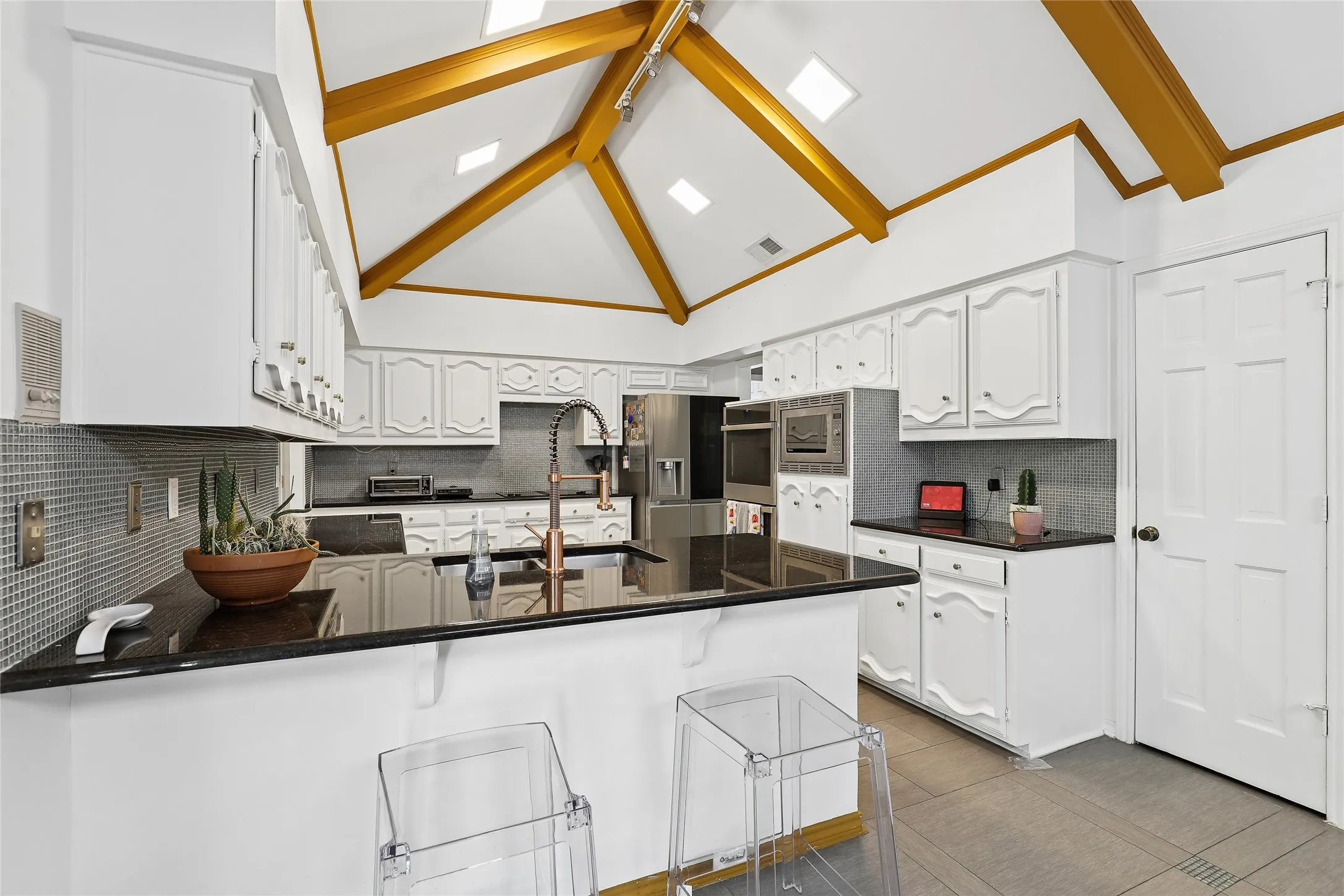 Vaulted ceiling with exposed beams, white cabinetry, dark
granite countertops, mosaic tile backsplash, and built-in stainless-steel appliances