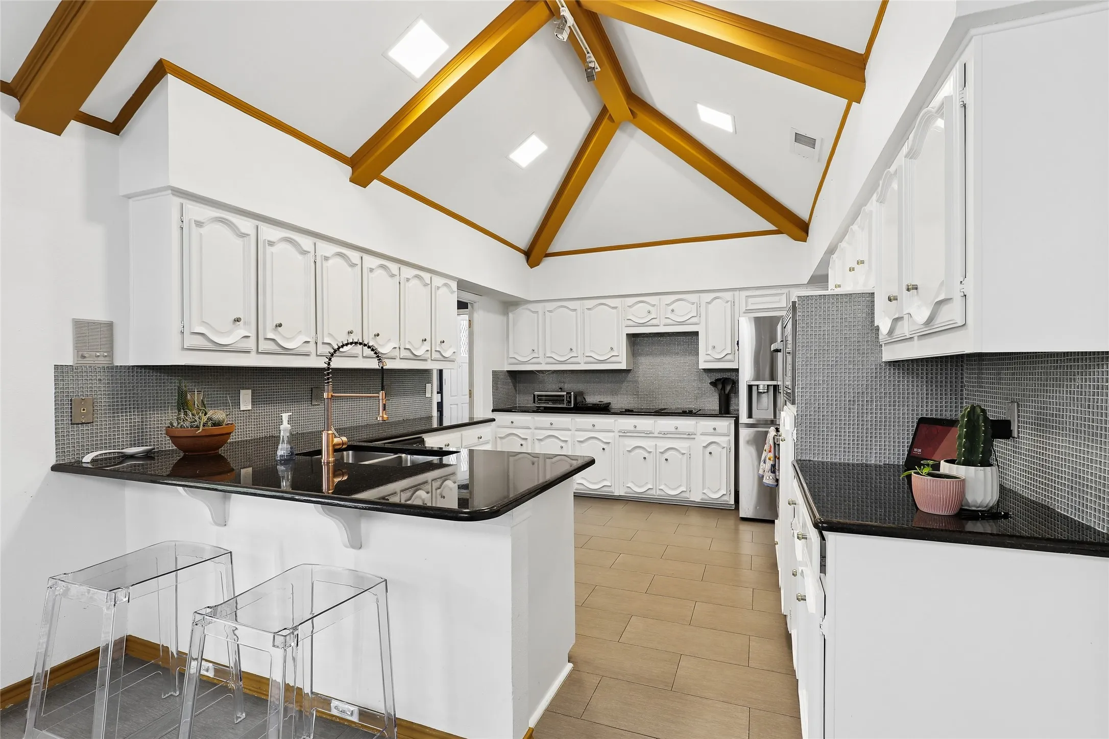 Vaulted ceiling with exposed wood beams, white cabinetry, dark countertops, gray mosaic tile backsplash, and light wood-finish flooring
