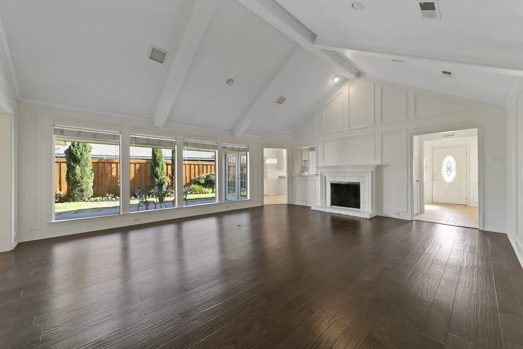 Dark wood-finish flooring, vaulted ceiling with exposed beams, white paneling accents, classic white fireplace, and a wall of windows overlooking a private yard