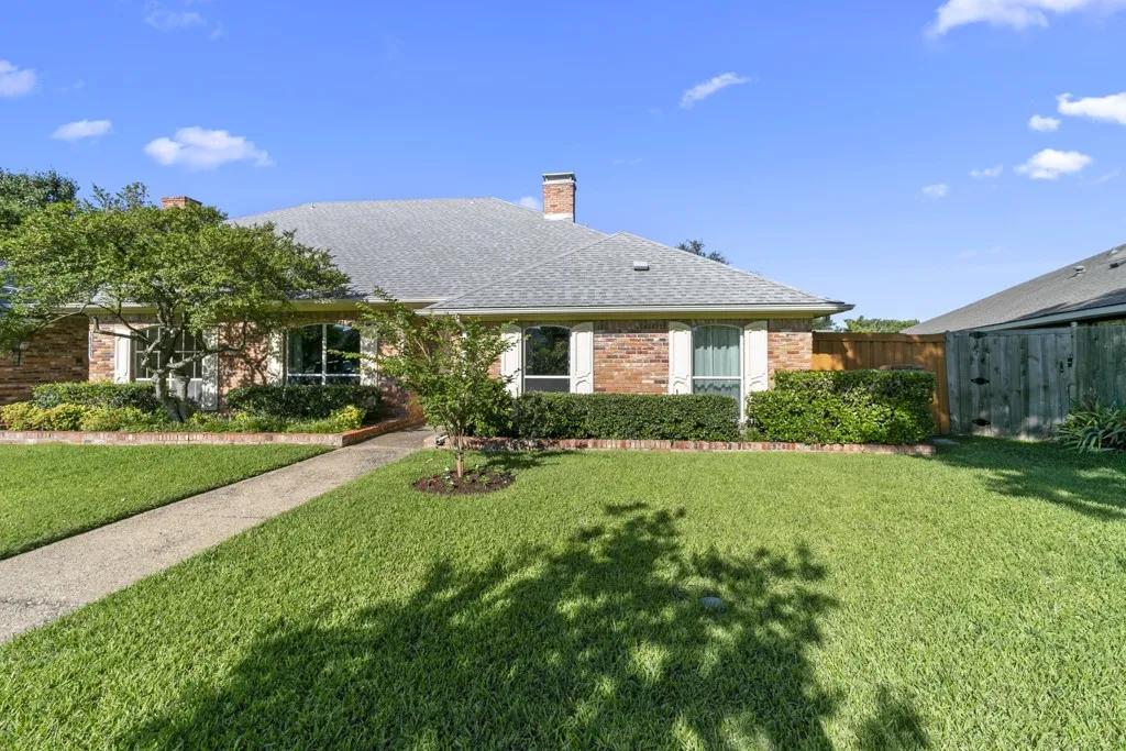 Brick exterior with a gray shingled roof, featuring white window shutters, mature landscaping, and a concrete pathway
