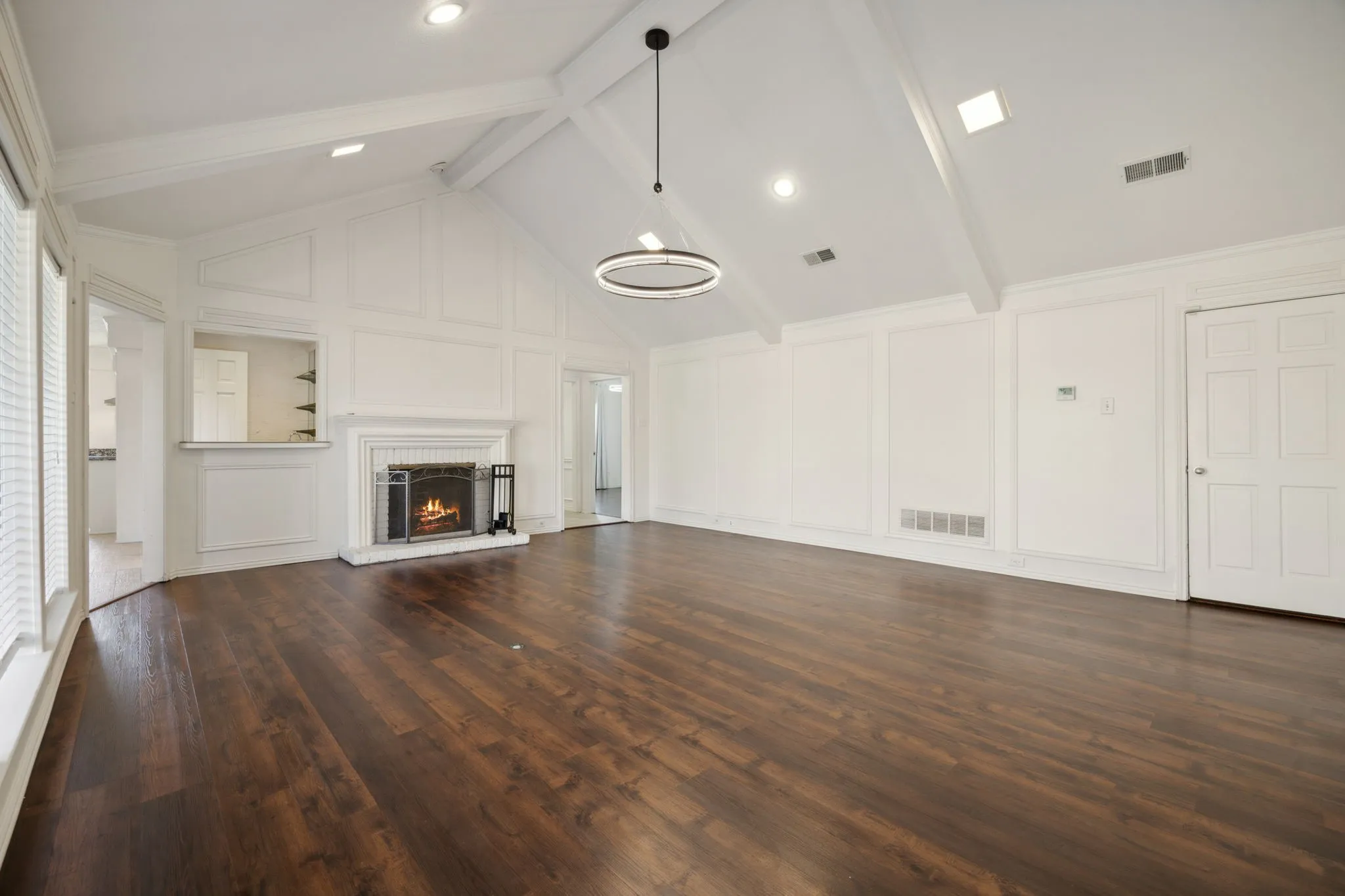 Spacious room with wood-finish flooring, a vaulted ceiling with exposed beams, and a fireplace featuring a white brick surround