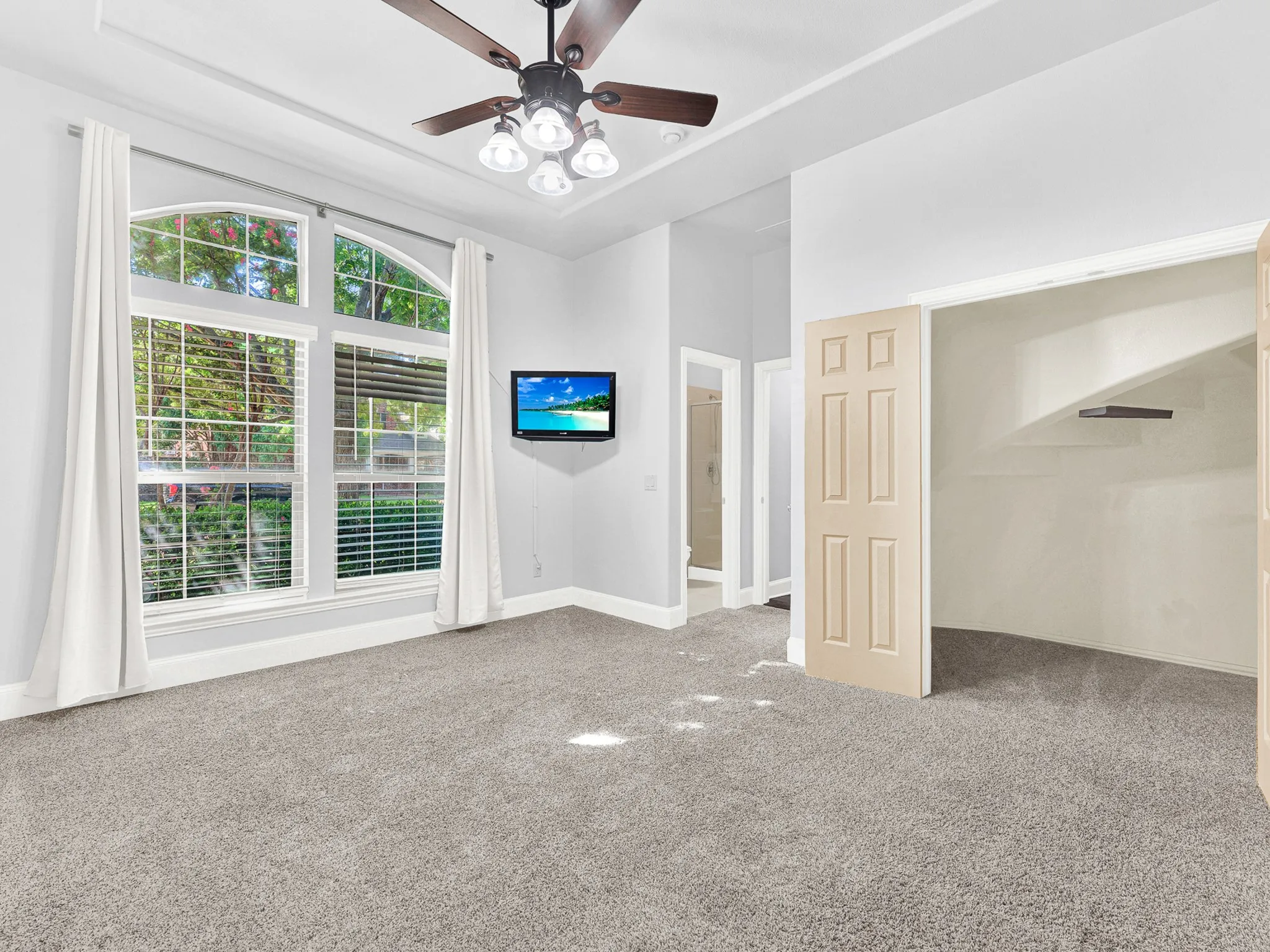 Arched window with blinds, ceiling fan with light fixture, tray ceiling, wall-mounted television, and carpeted flooring