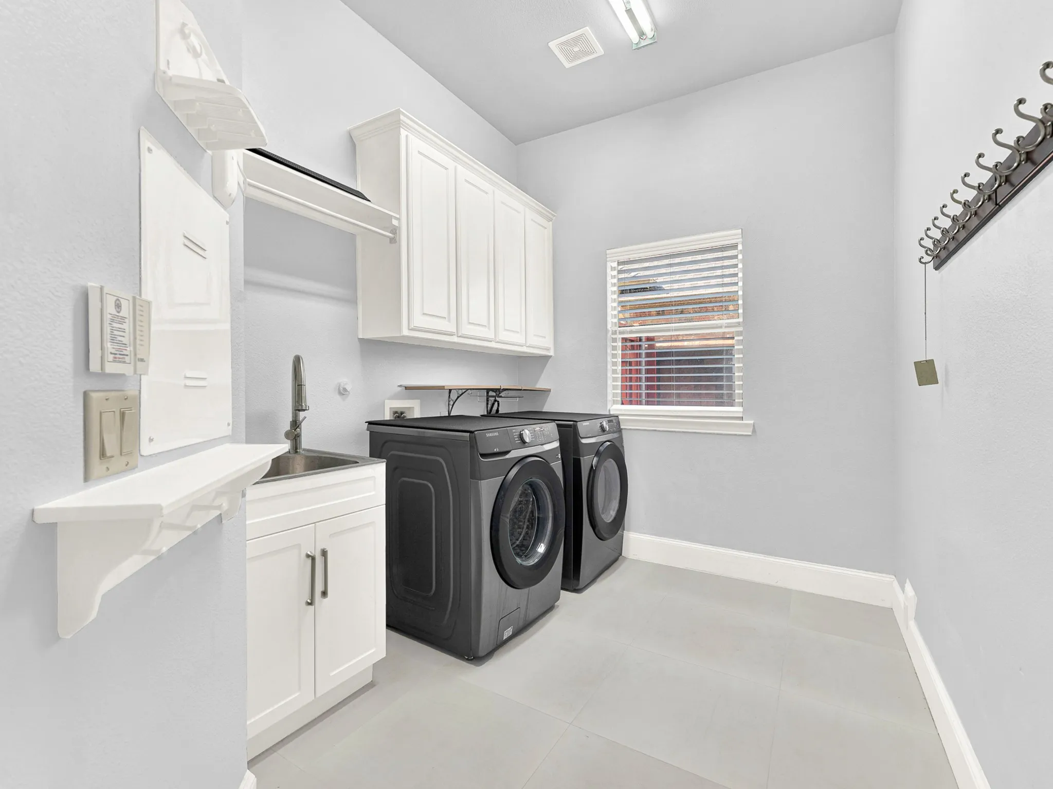 White shaker-style cabinetry, stainless steel sink, light-colored tile flooring, and a window with blinds