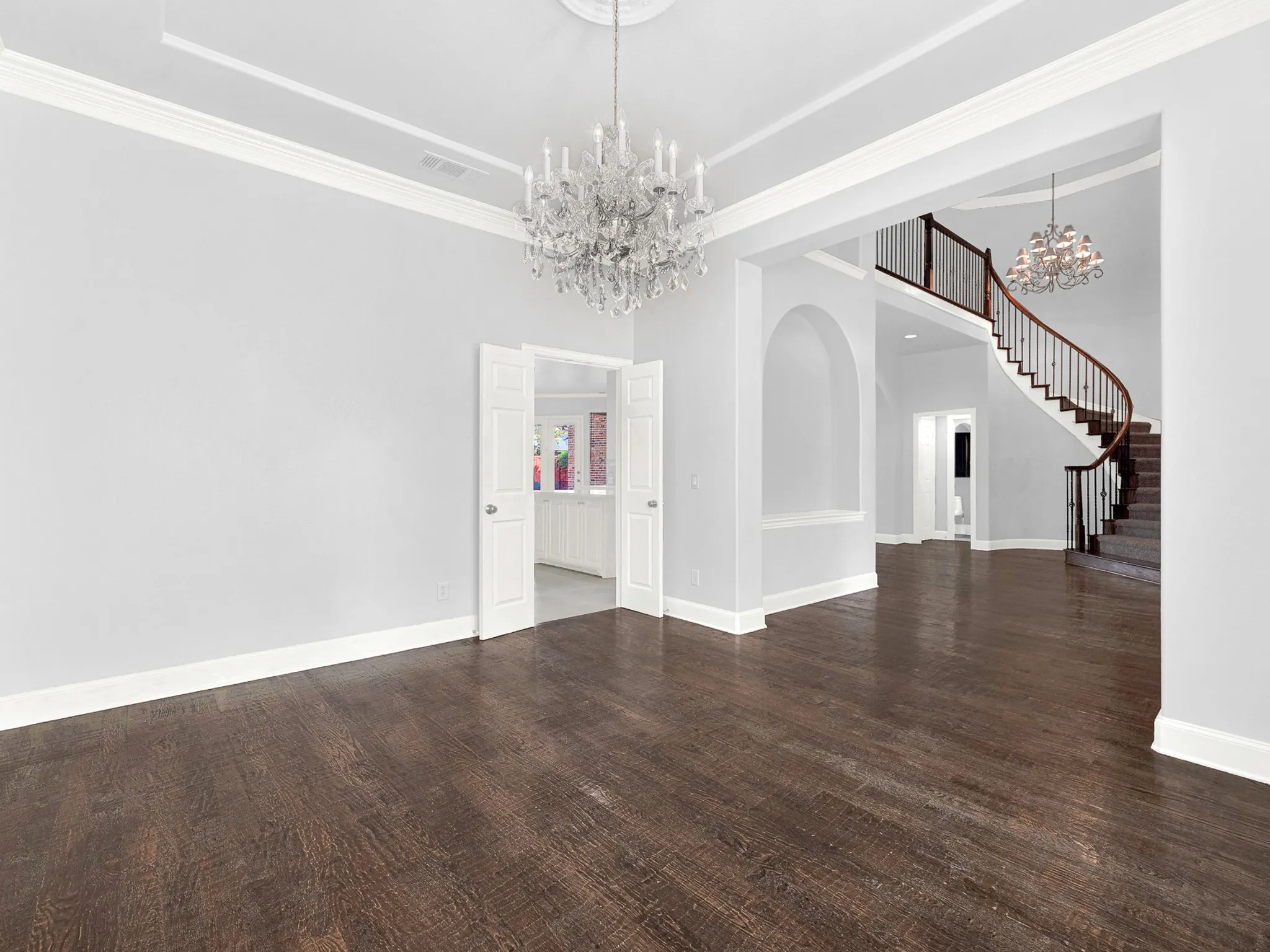 Dark wood-finish flooring, two crystal chandeliers, a grand curved staircase with wood railing, white crown molding, and an arched entryway