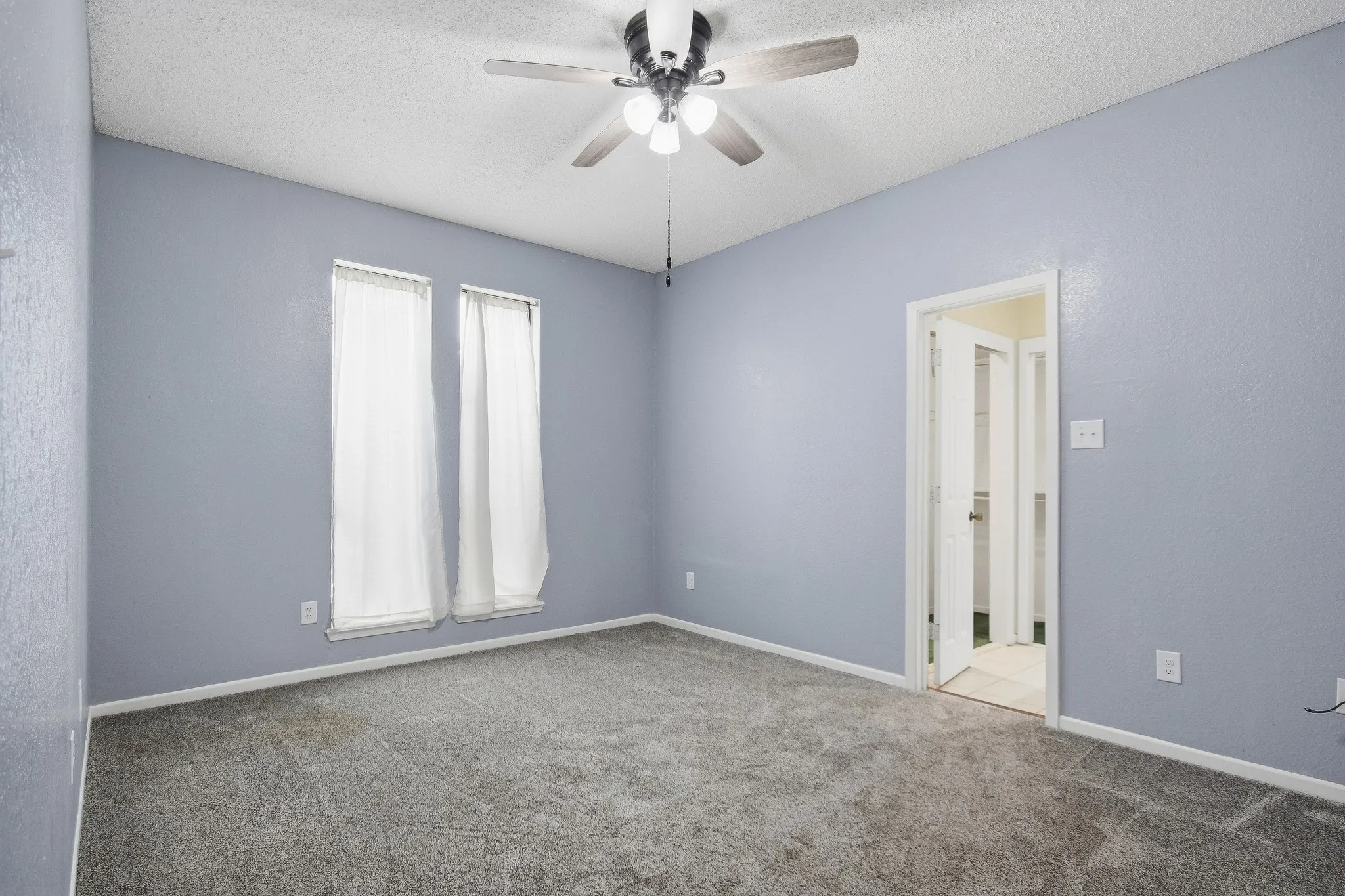 Gray carpeted room featuring two tall windows, a ceiling fan with integrated lighting, and a white paneled door
