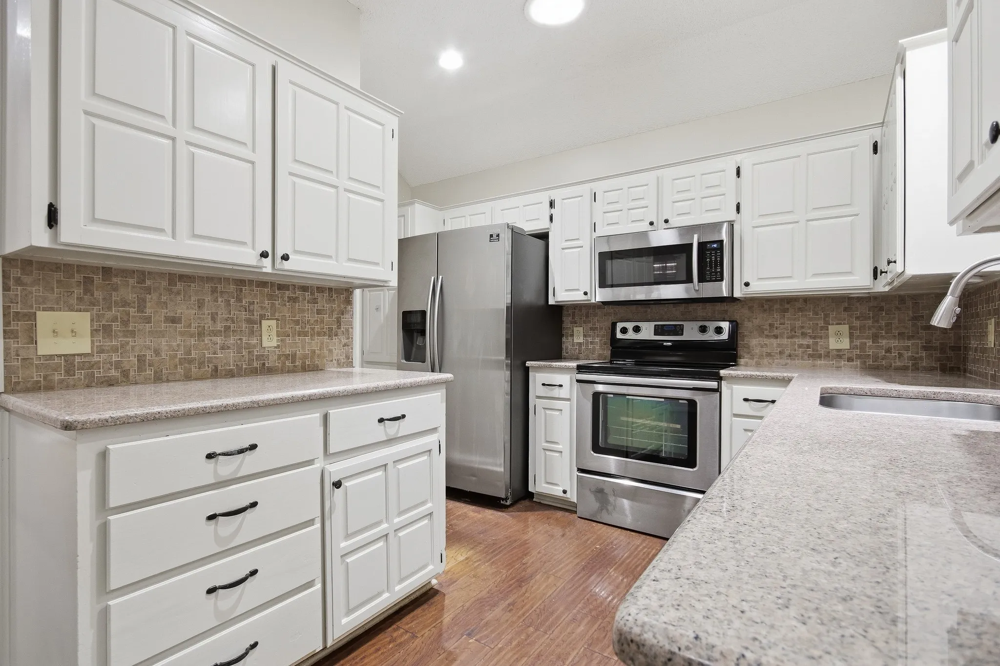 White paneled cabinetry, stainless steel appliances, speckled countertops, mosaic tile backsplash, and wood-finish flooring