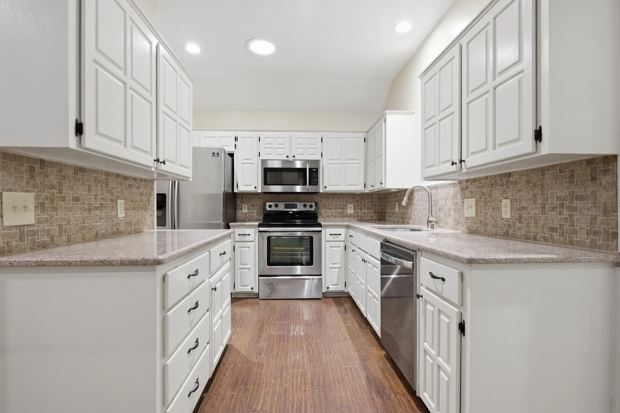 White cabinetry, stainless steel appliances, mosaic tile backsplash, speckled countertops, and wood-finish flooring