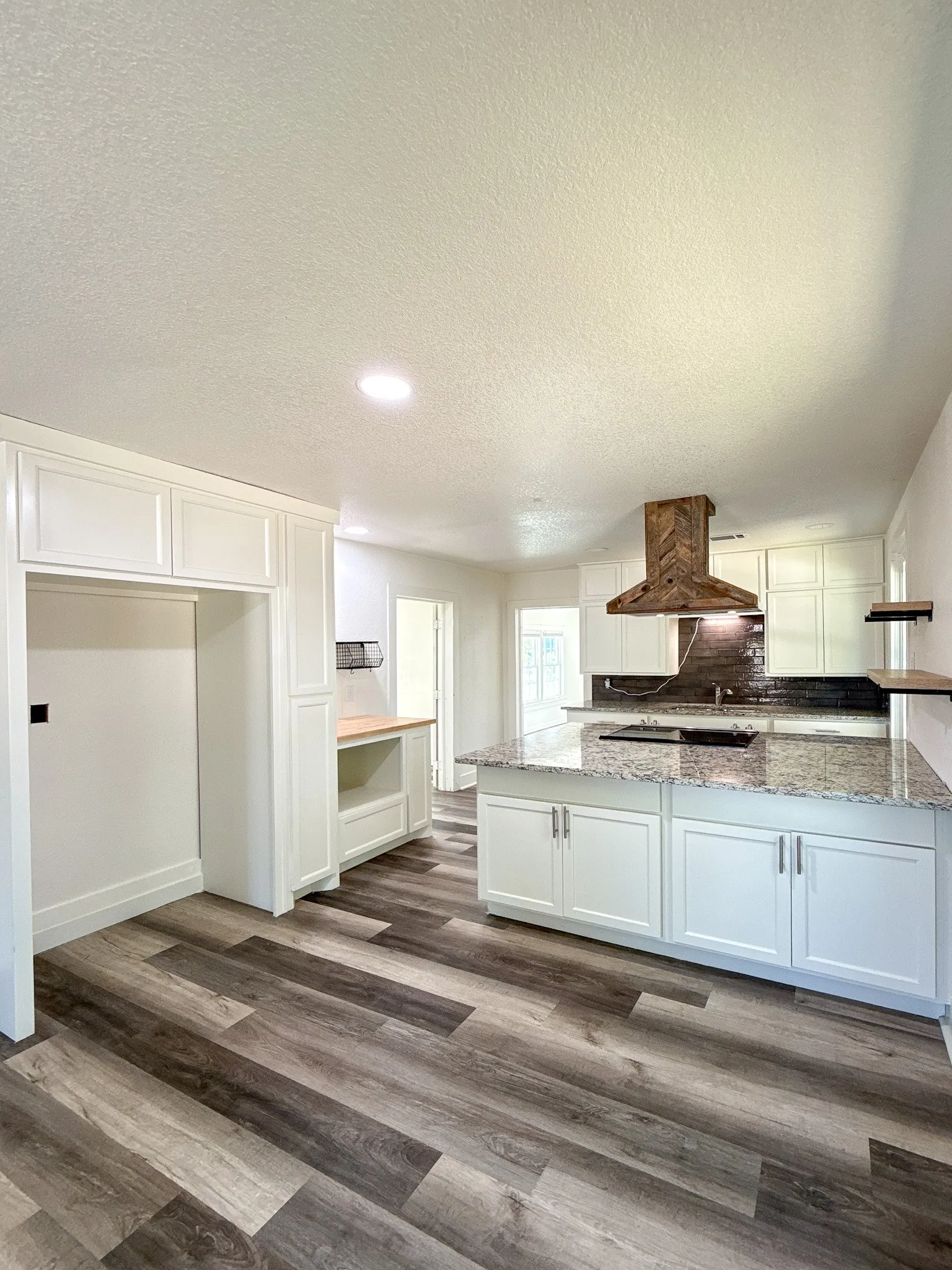 Kitchen featuring wood-finish flooring, white cabinetry, a central island with a light-toned countertop, a dark tile backsplash, and a wood-finish range hood