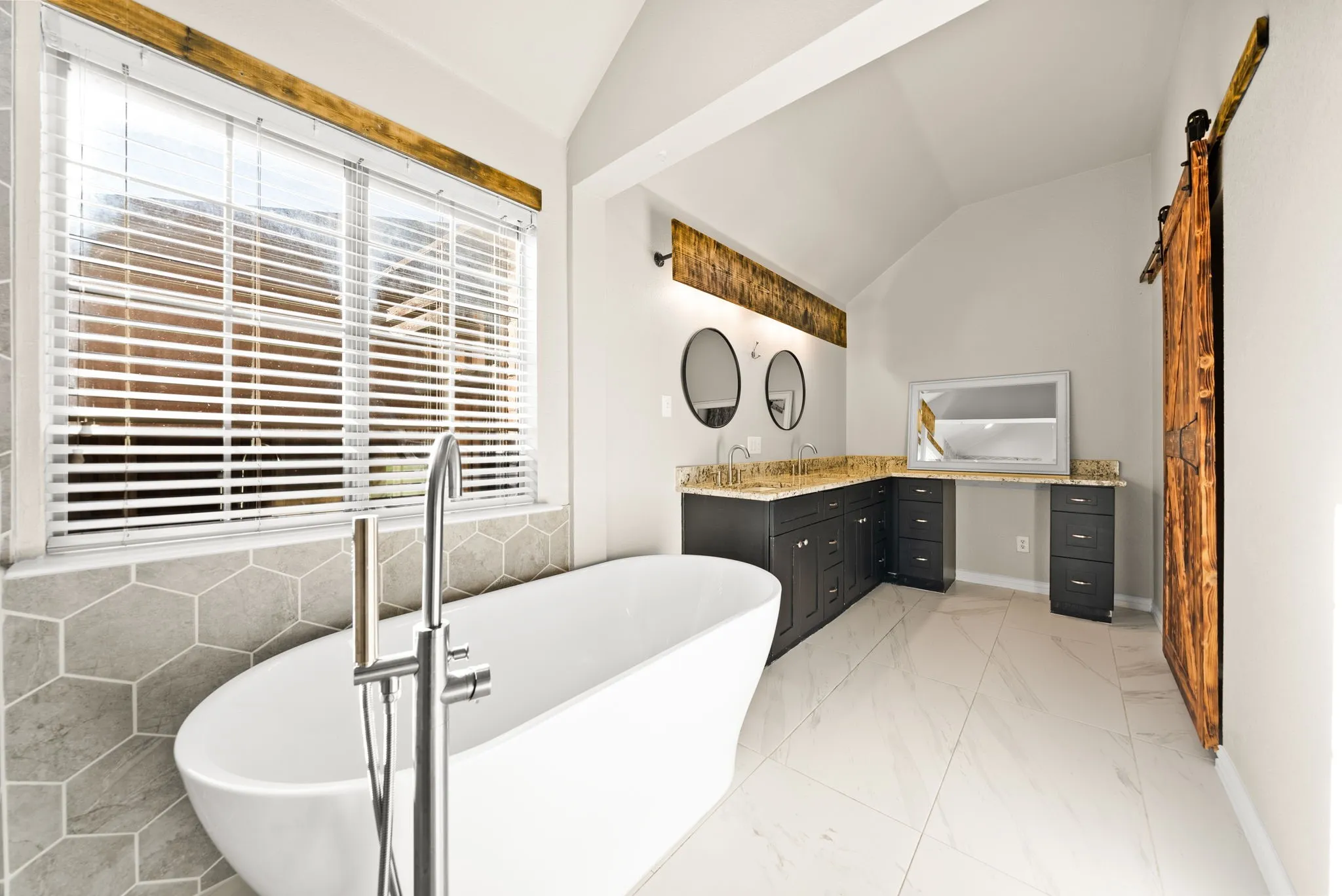Spa-inspired primary bathroom featuring a freestanding soaking tub with floor-mounted filler, dual vanity with black cabinetry and wood-finish countertops, vessel sinks, polished nickel hardware, and large-format tile flooring
