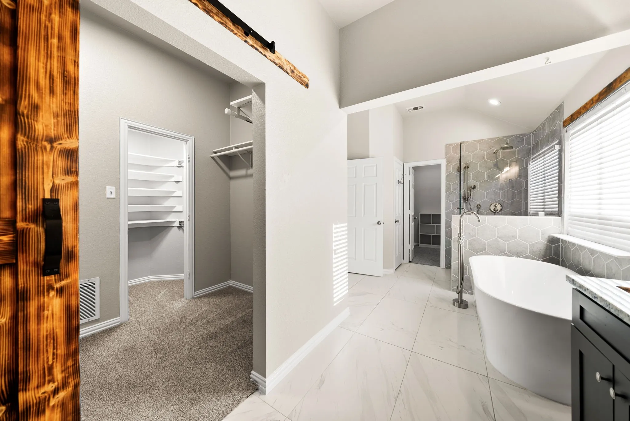 Freestanding bathtub with floor-mounted faucet, hexagon tile shower surround, wood-finish barn door, and walk-in closet with built-in shelving
