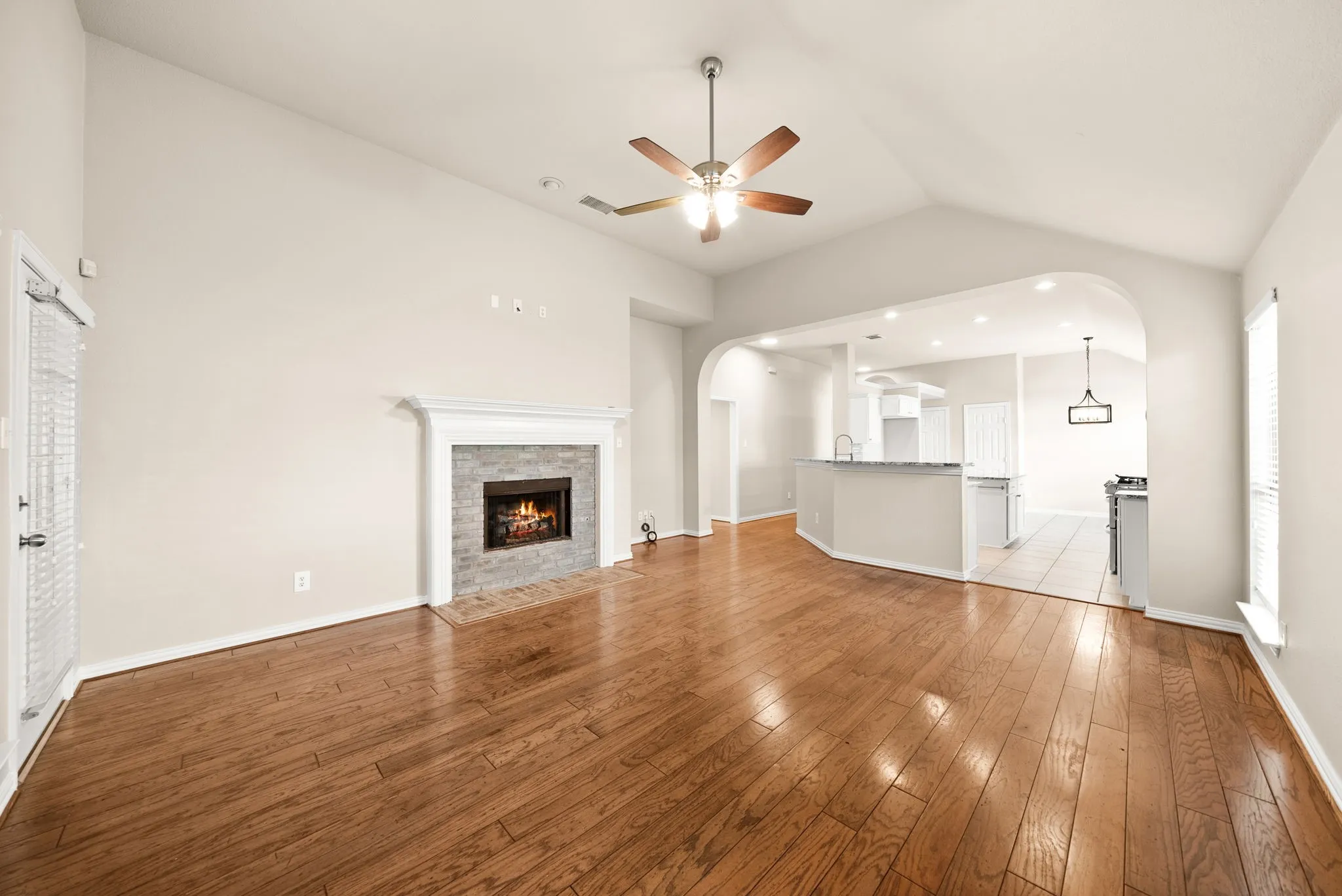 Vaulted ceiling with wood-finish ceiling fan, wood-finish flooring, stone-faced fireplace with white mantel, kitchen pass-through with breakfast bar, and recessed lighting