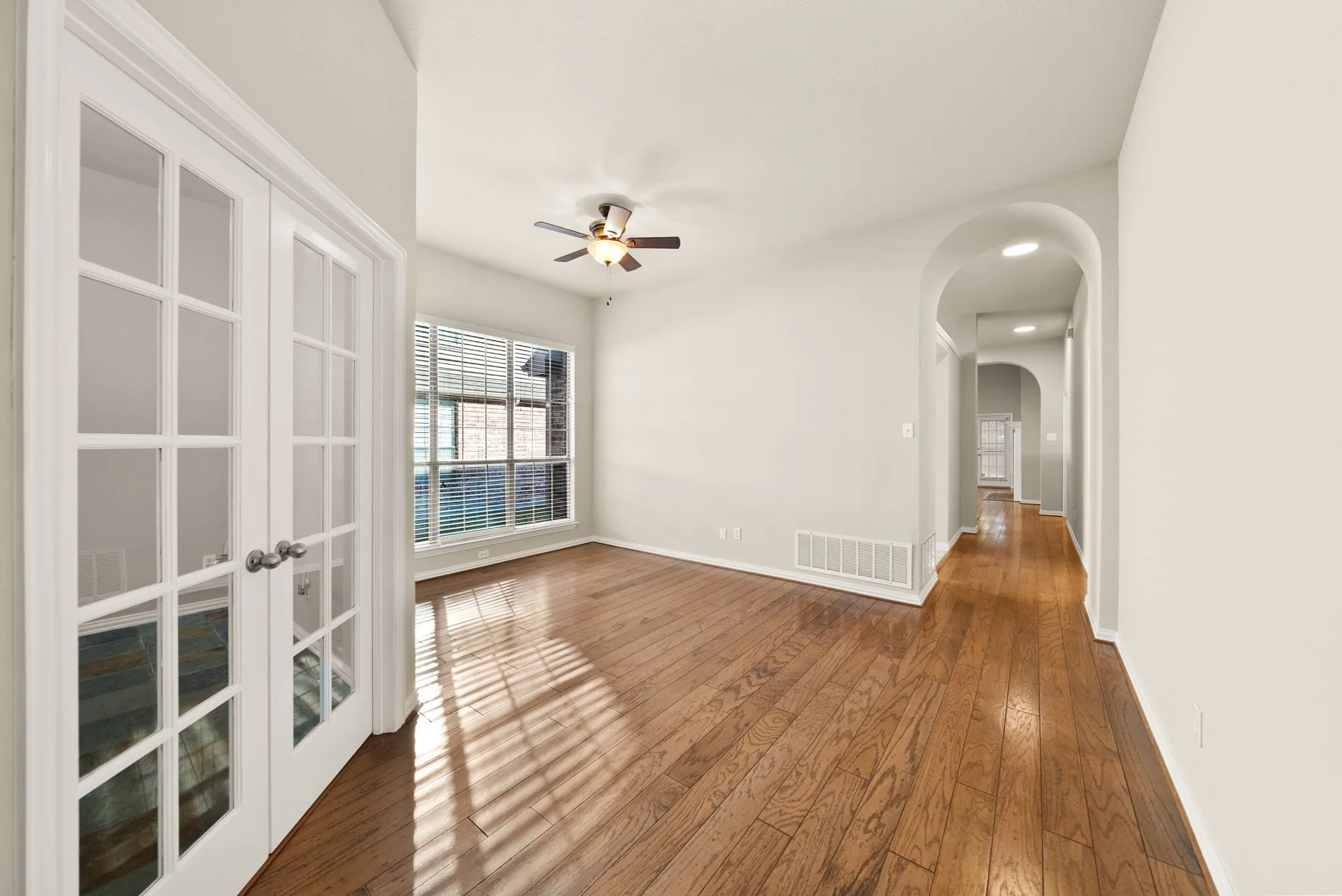 Hardwood flooring throughout, French doors with glass panels, large window with blinds, ceiling fan with light fixture, and arched hallway entry