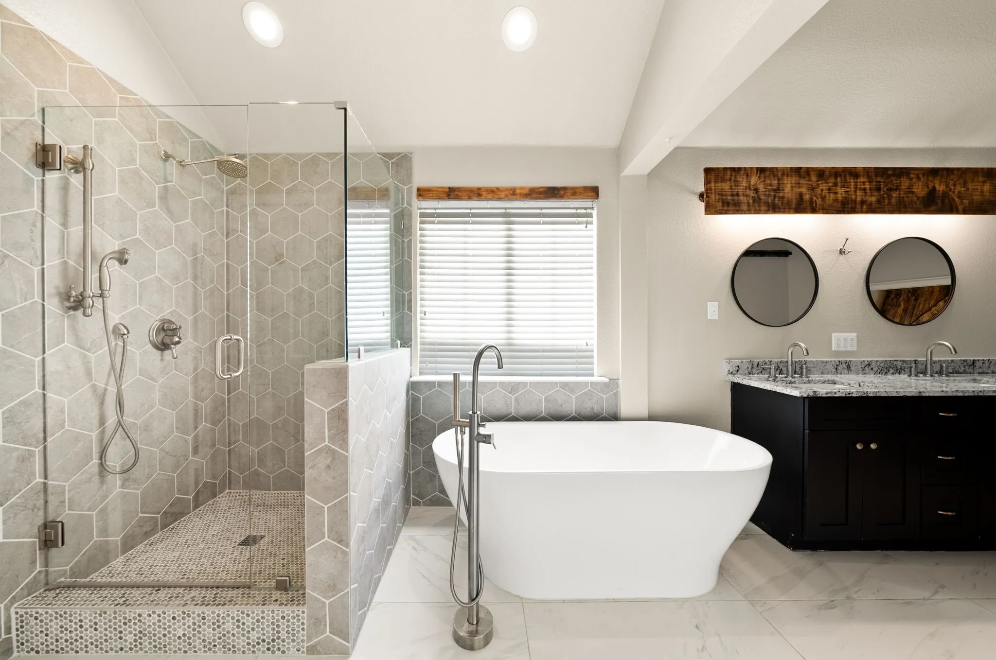 Freestanding soaking tub, walk-in shower with glass enclosure, dual vanity with undermount sinks, wood-finish beam with integrated lighting, and hexagonal tile accents