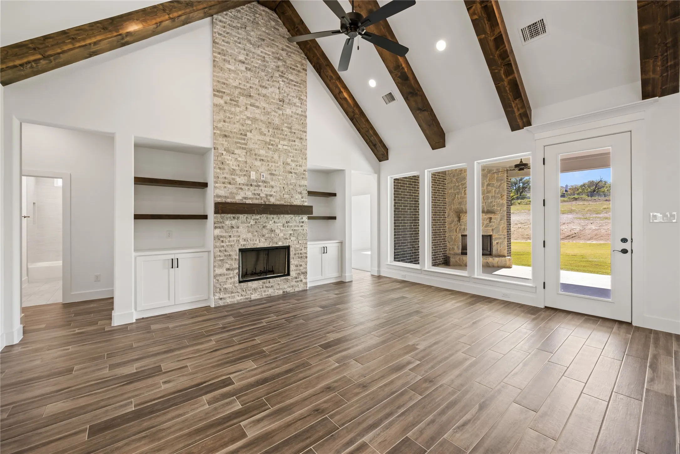 Vaulted ceilings with exposed wood beams, a floor-to-ceiling stone fireplace, built-in cabinetry with wood-finish shelving, wood-finish tile flooring, and multiple large windows with exterior access
