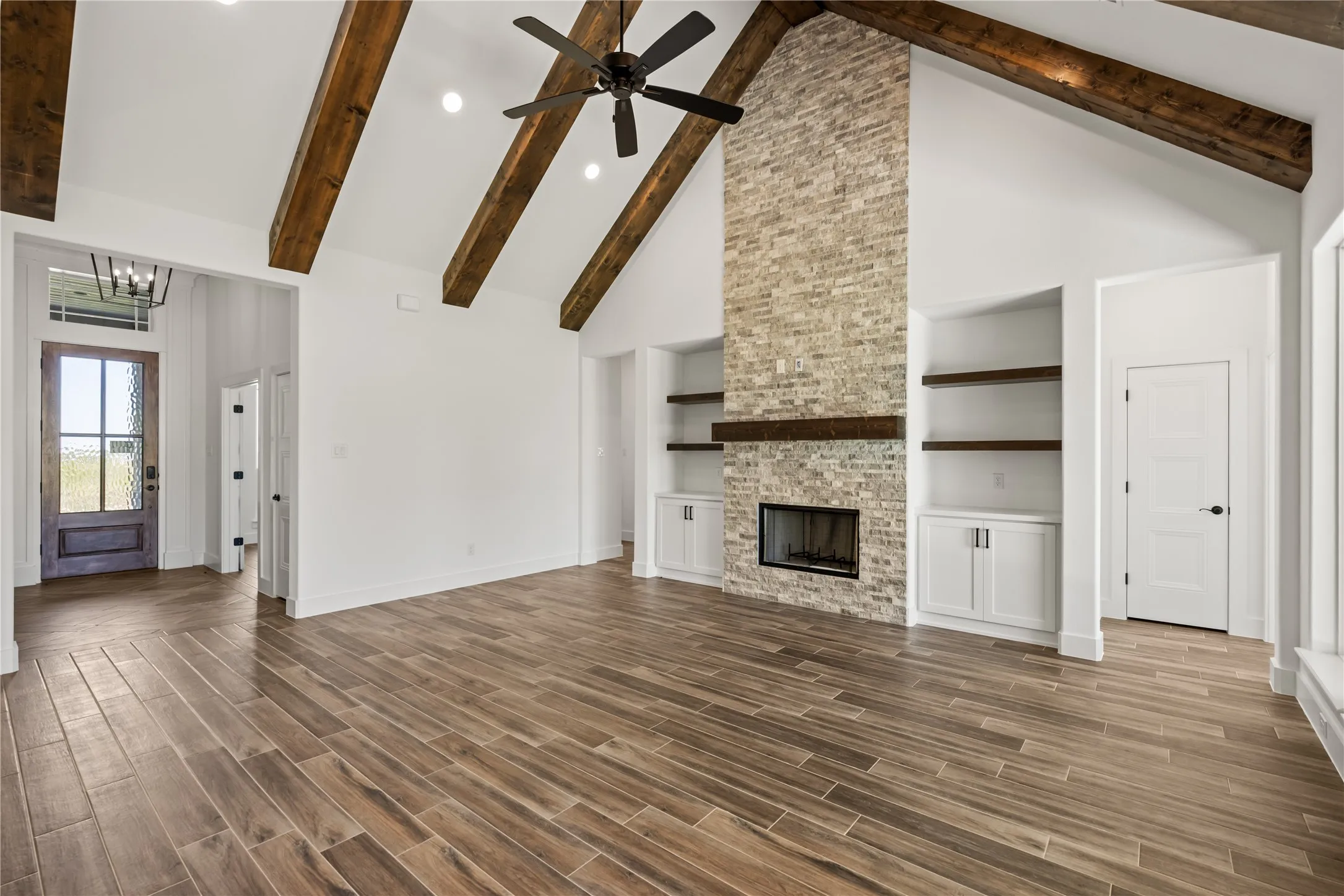 Vaulted ceiling with exposed wood beams, wood-finish flooring, stone fireplace with a wood mantle, recessed lighting, and built-in cabinetry with open shelving