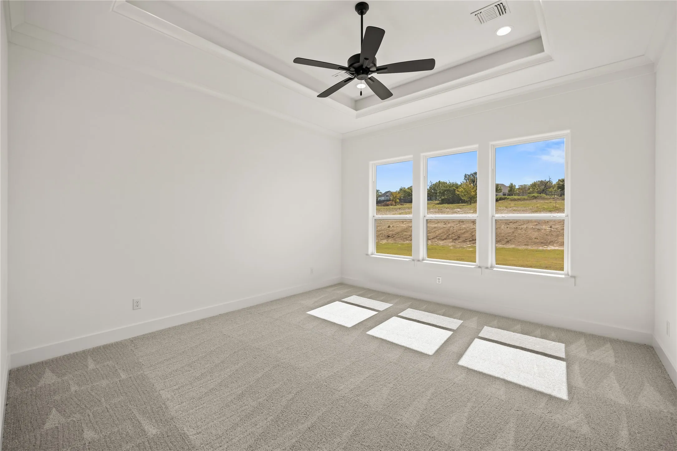 Recessed ceiling with crown molding, three large windows, ceiling fan, and light-toned carpet flooring