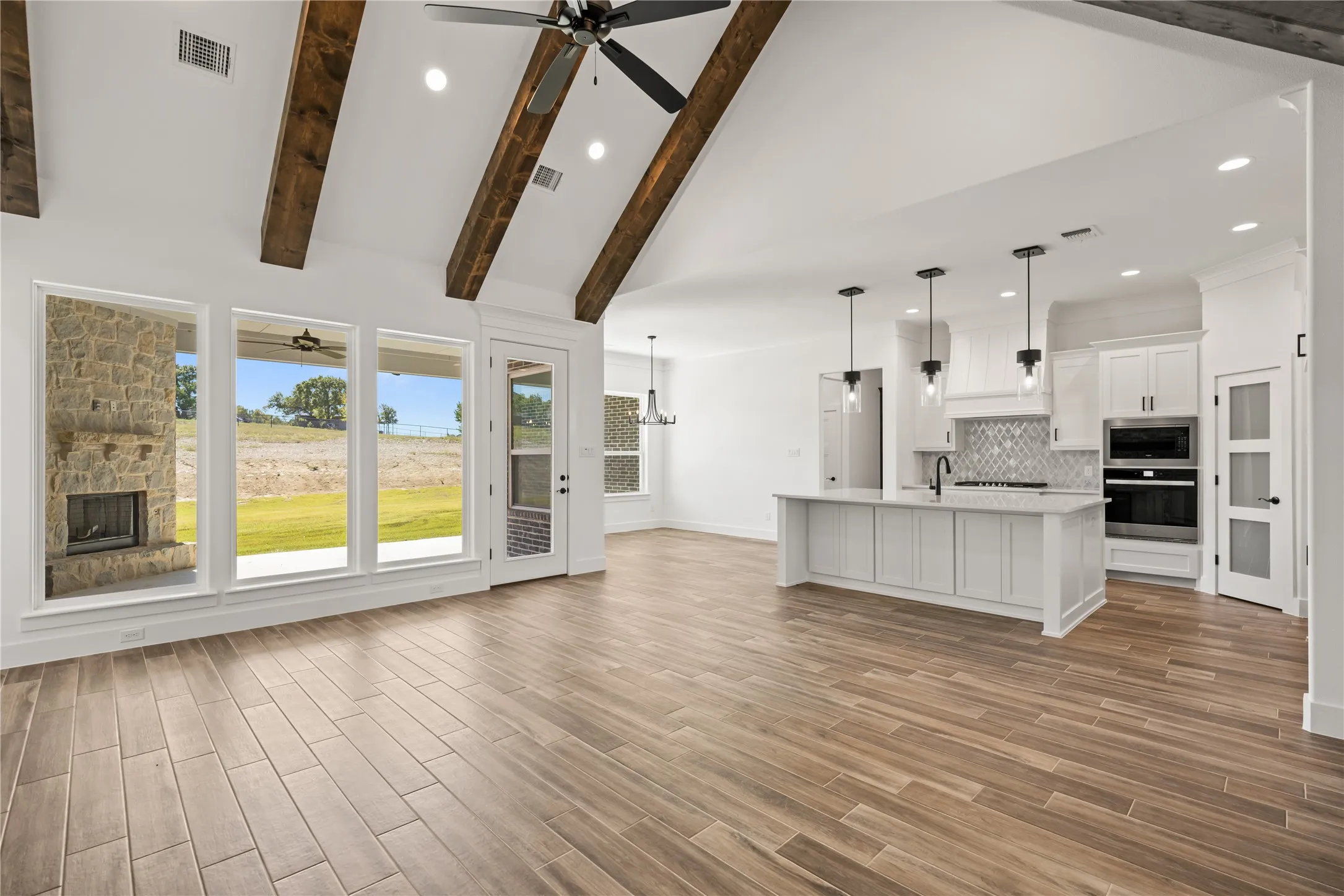 Vaulted ceiling with exposed wood beams, stone fireplace, wood-finish flooring, white cabinetry, and a kitchen island with pendant lighting