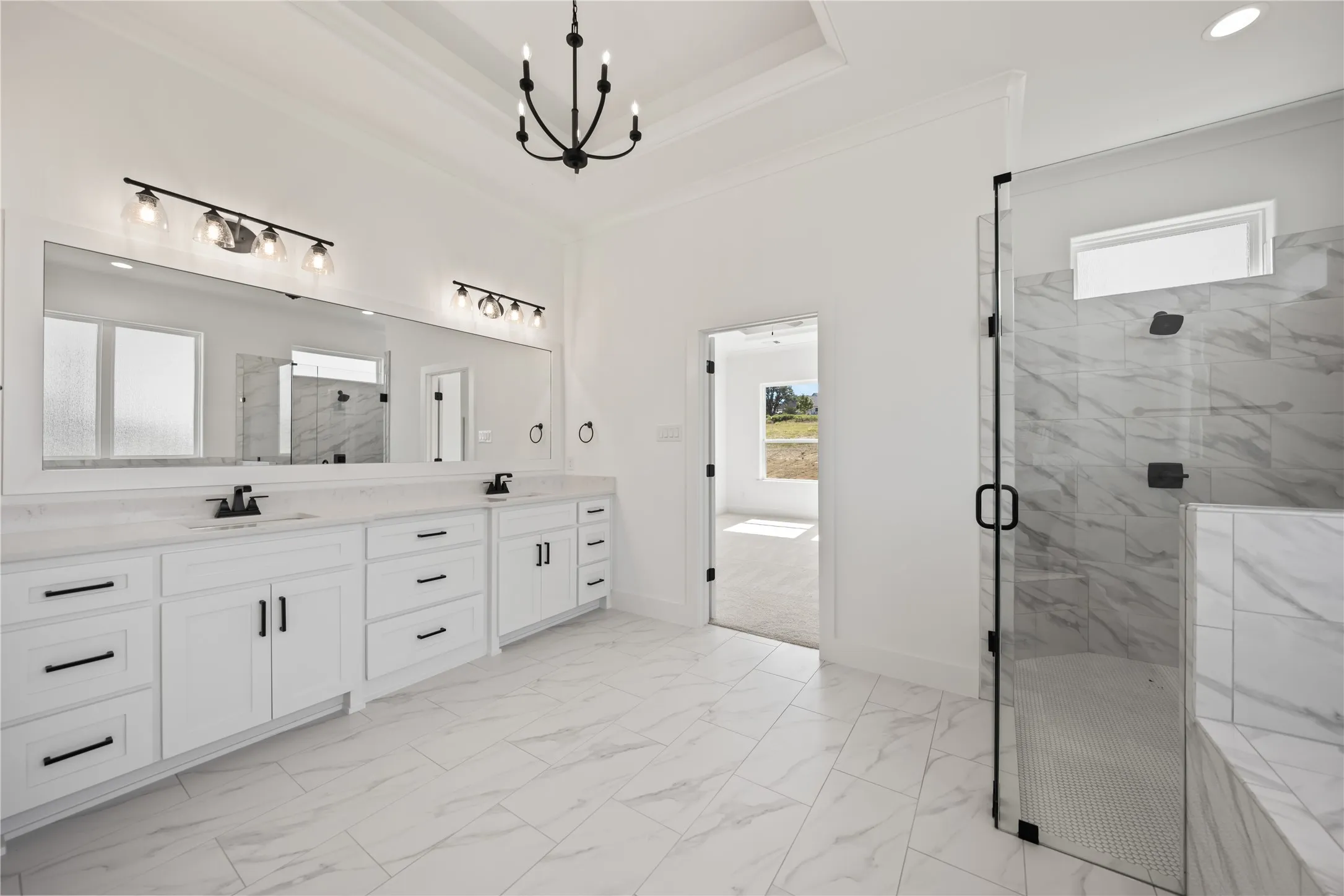 Dual vanity with white cabinetry and dark hardware, large framed mirror, walk-in shower with glass enclosure, white tile flooring, and tray ceiling