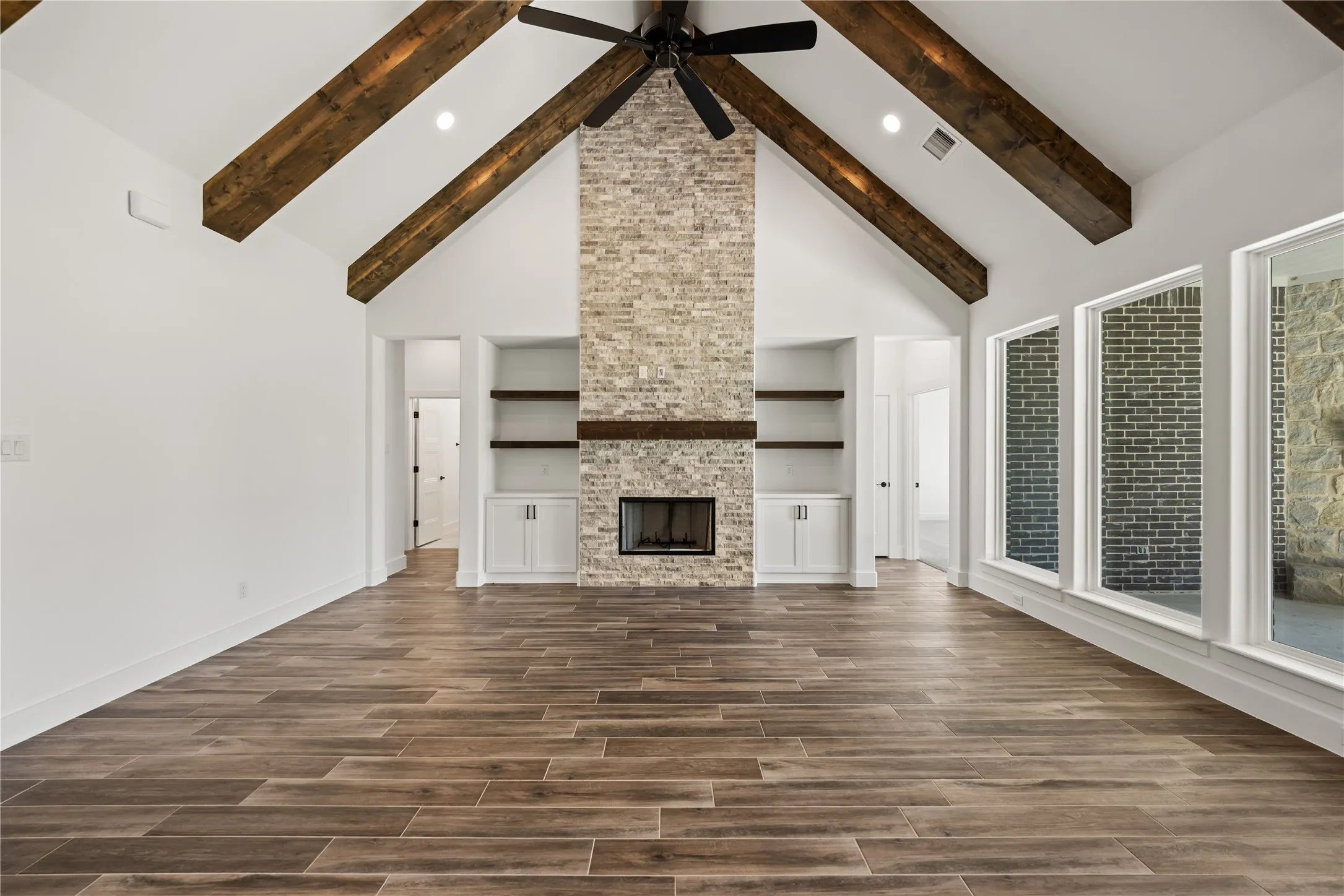 Vaulted ceiling with exposed wood beams, brick fireplace with wood mantle, built-in shelving and cabinetry, wood-finish flooring, and multiple large windows