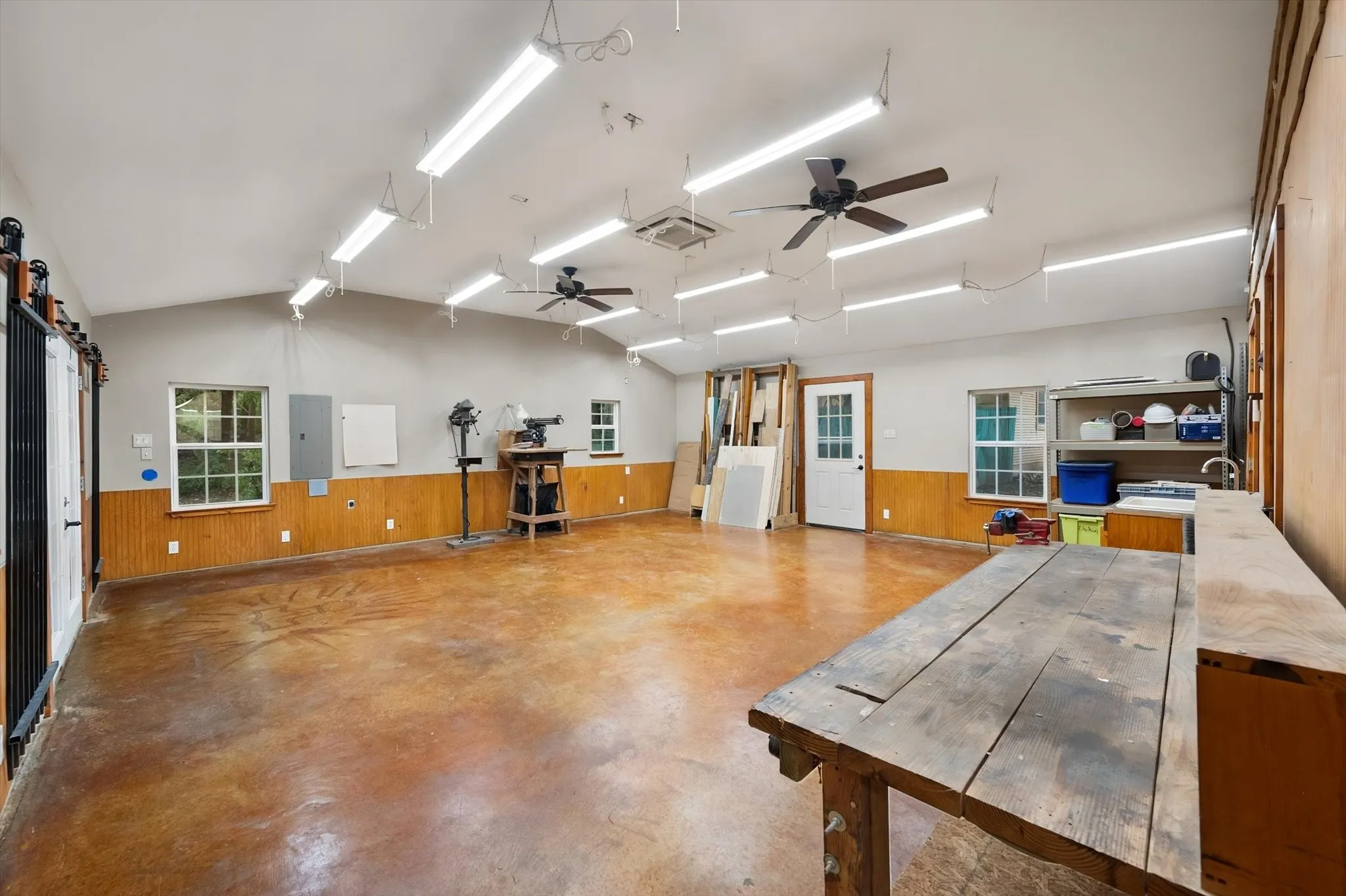 Vaulted ceiling with multiple linear fluorescent light fixtures, two ceiling fans, and concrete-finish flooring