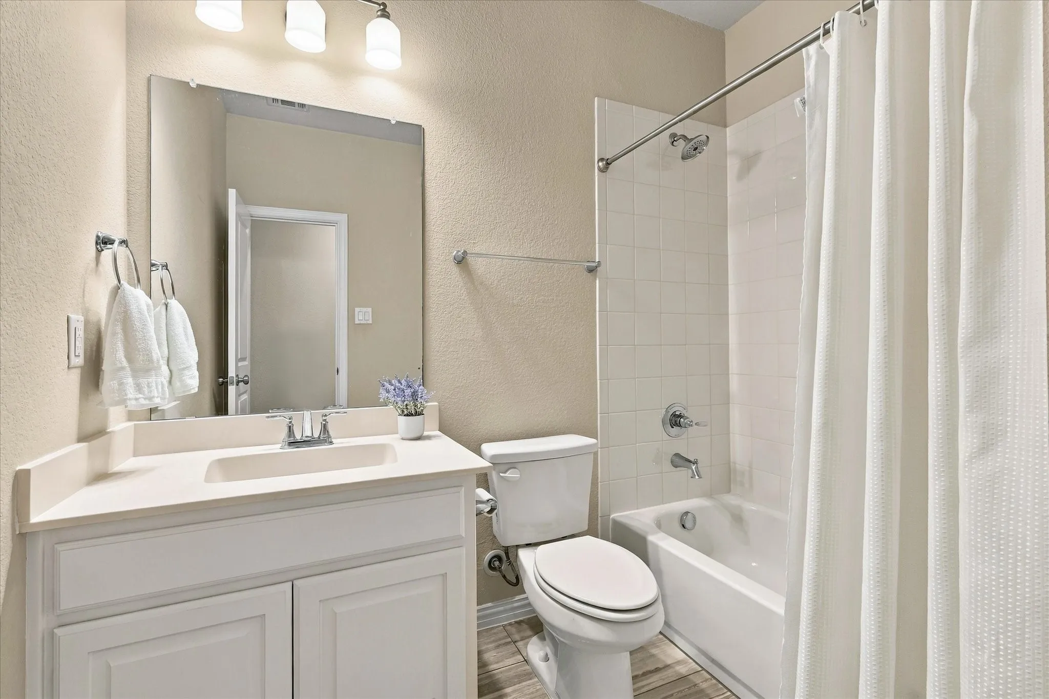 White vanity with integrated sink, rectangular mirror, three-light vanity fixture, white tiled tub/shower combination, and wood-finish flooring