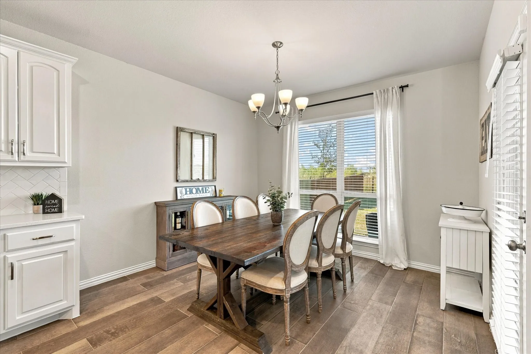 Wood-finish flooring, white cabinetry with hardware, herringbone tile backsplash, large window with blinds, and a chandelier lighting fixture