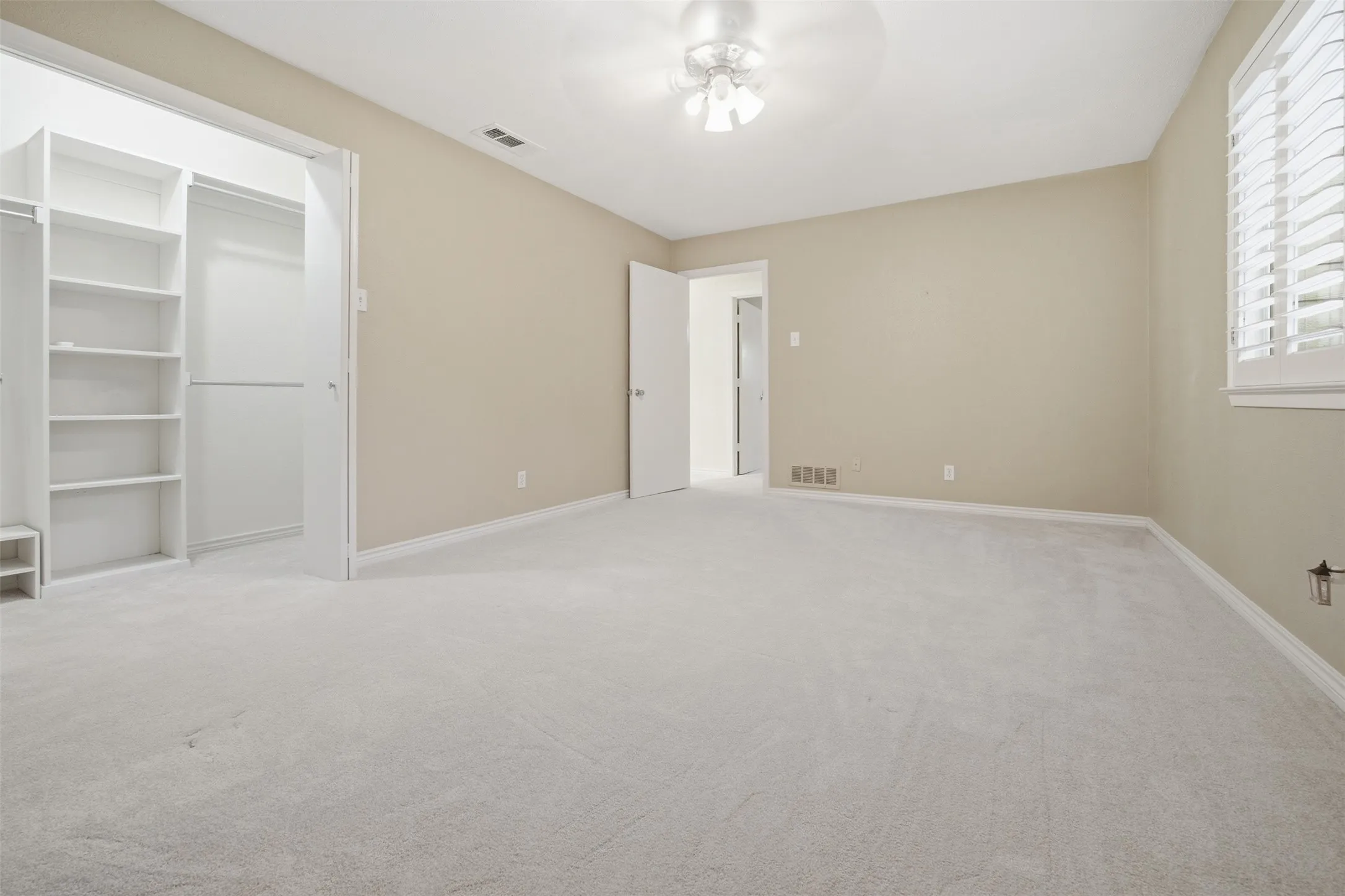 Wall-to-wall carpeting, window with plantation shutters, ceiling fan with light fixture, and built-in closet shelving
