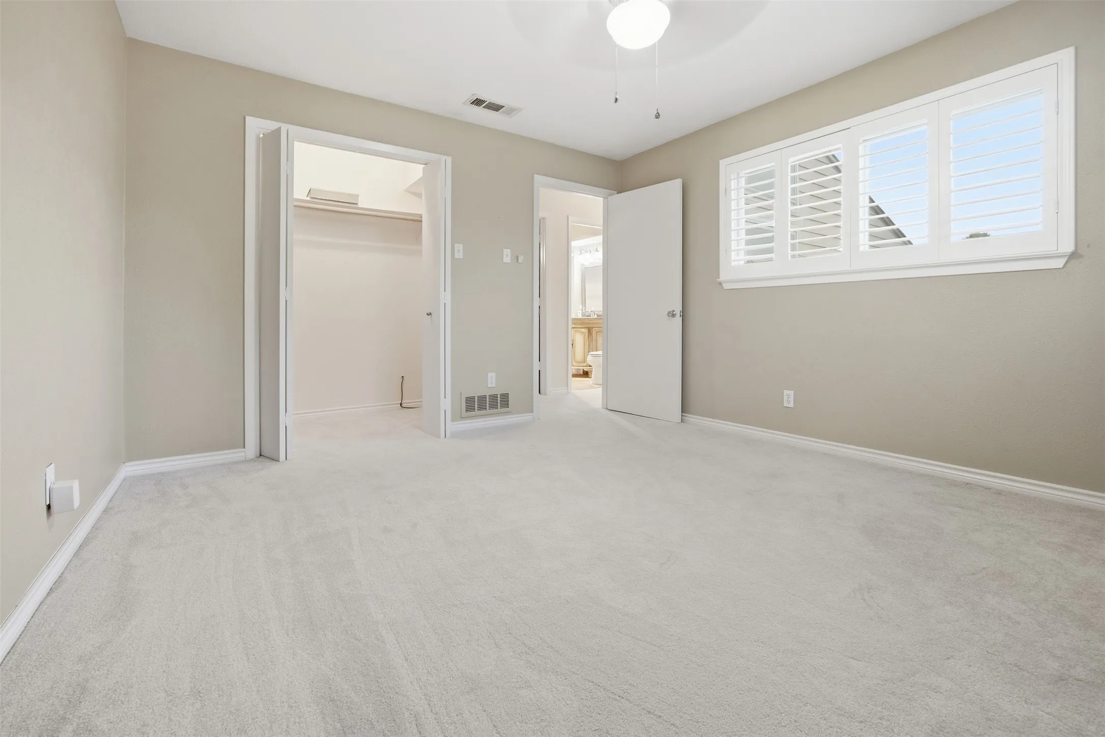 Light neutral wall color, plush light-toned carpeting, window with plantation shutters, ceiling fan with integrated lighting, and built-in closet shelving